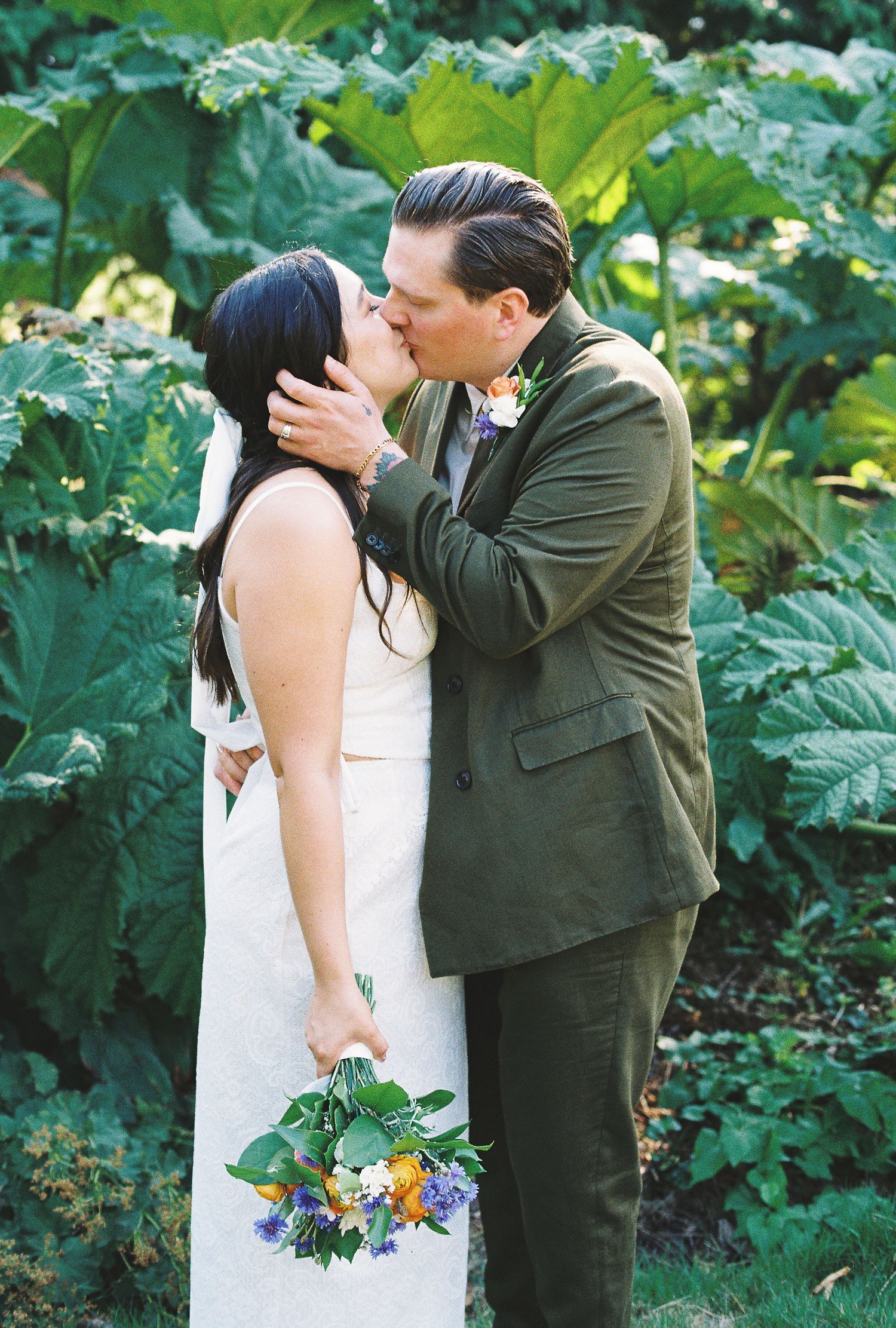 Bride and groom sharing first kiss surrounded by lush green tropical plants at wedding.