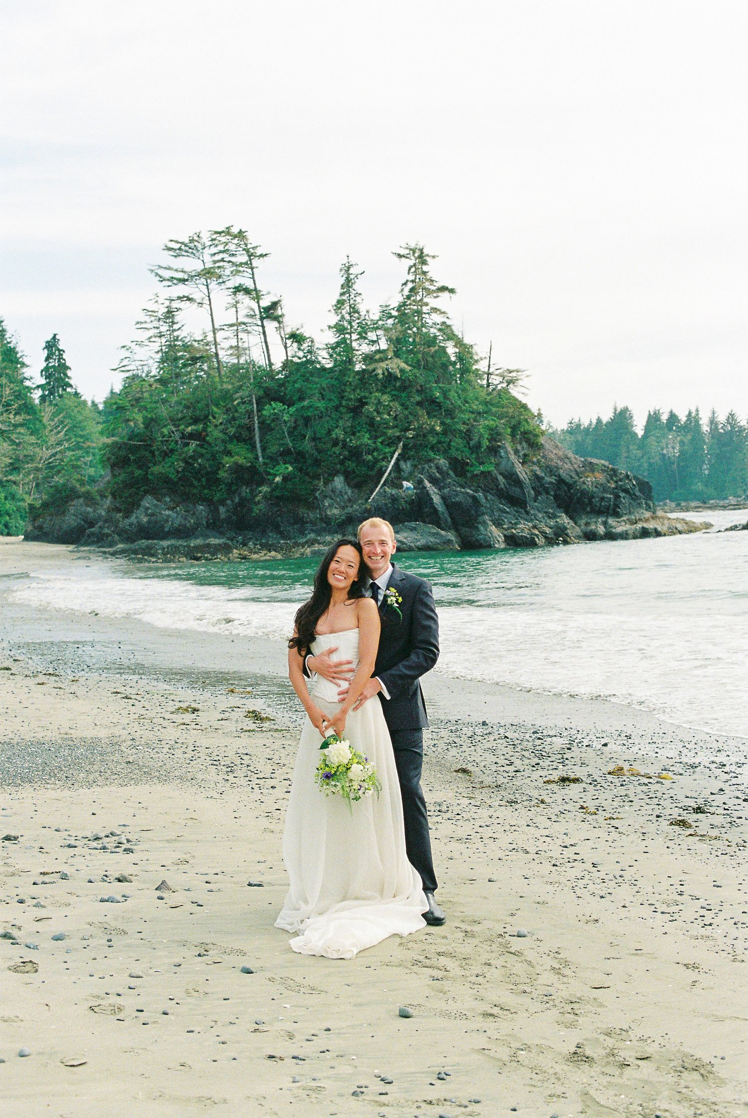 Bride and groom posing on rocky beach with lush forested island and ocean in background.