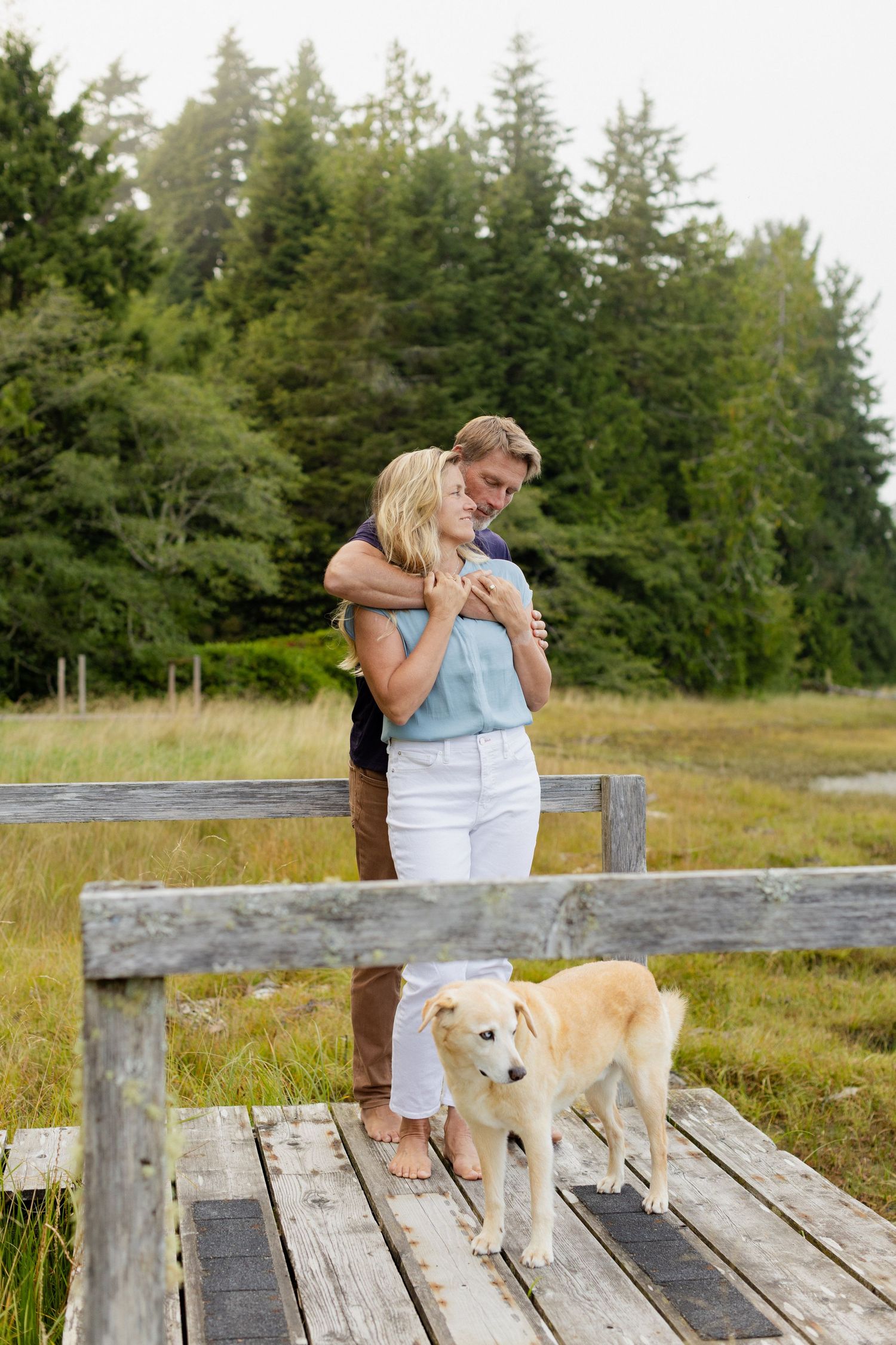 A couple embraces on a wooden dock while their golden dog stands nearby with evergreen trees behind them.