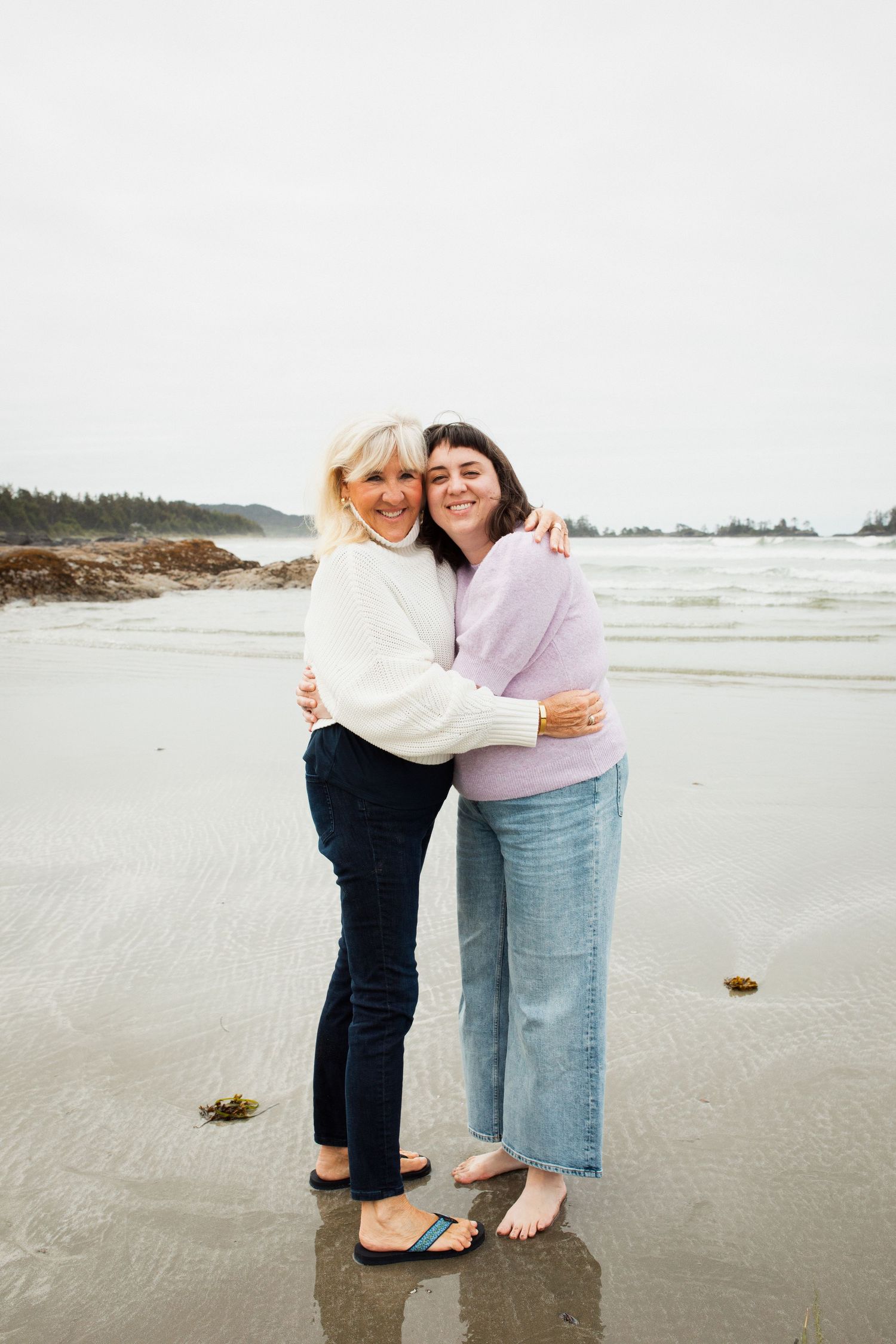 Two women embrace and smile together on a sandy beach with ocean waves in the background.