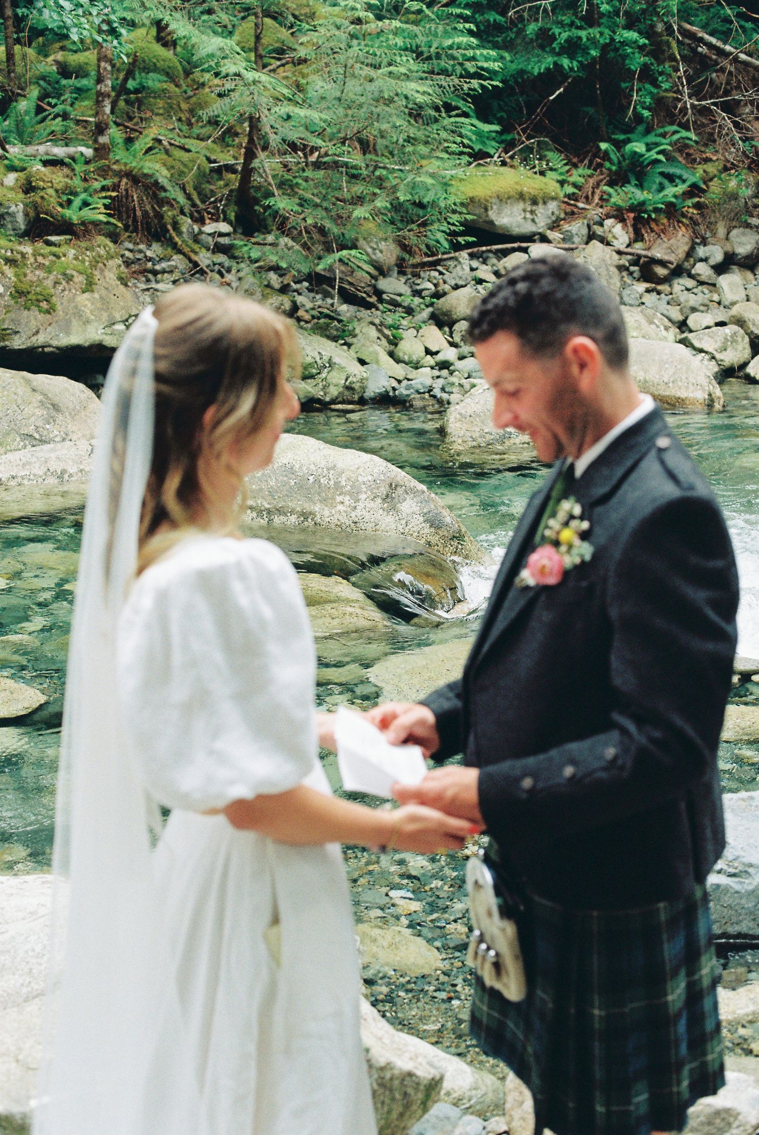 Groom in tartan kilt reads vows to bride in white dress beside a lush forest creek.
