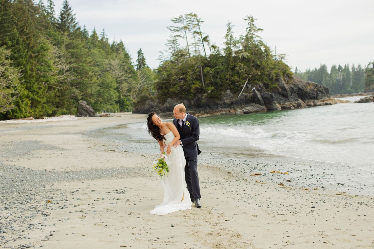 Bride and groom share a romantic embrace on a sandy beach surrounded by lush Pacific Northwest forest.