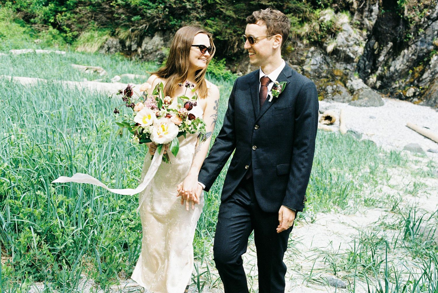 Bride in vintage gown with sunglasses holding lush bouquet walking with groom along a coastal path.