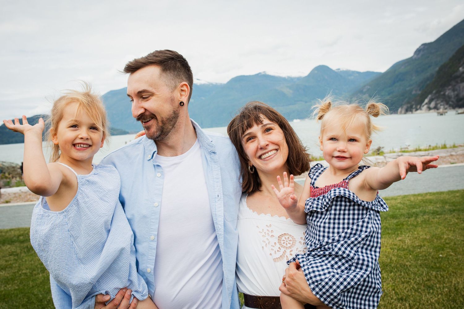 Happy family of four smiling outdoors with mountain lake backdrop, parents holding two young daughters.