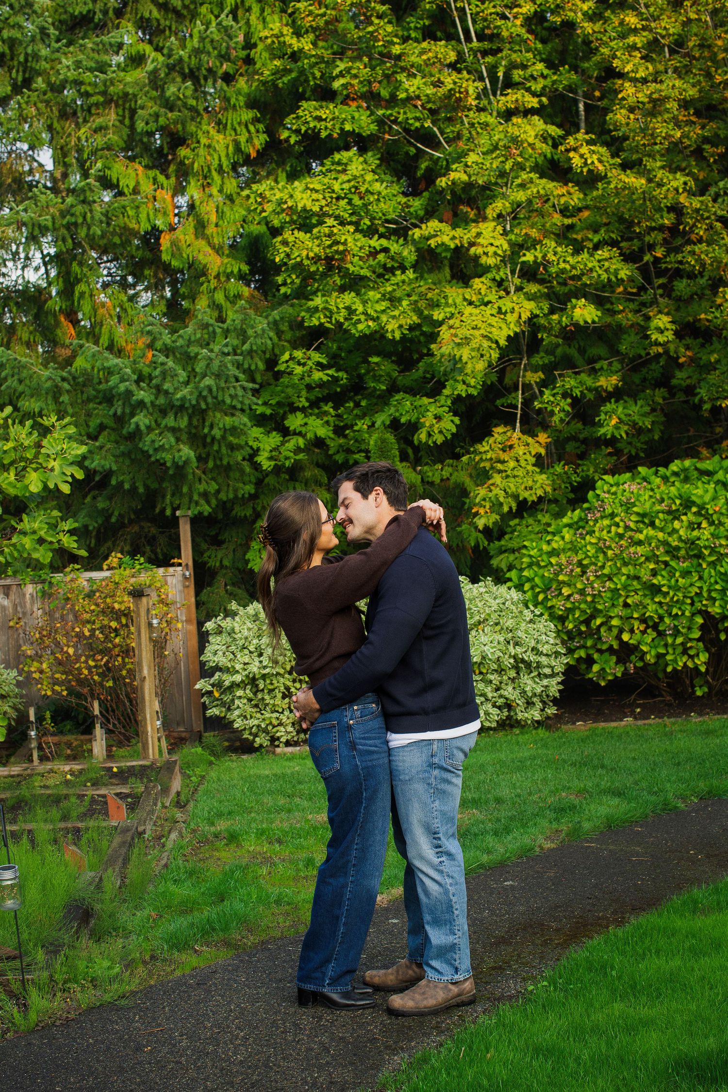 Couple sharing a tender kiss while embracing on a garden path surrounded by lush green trees.