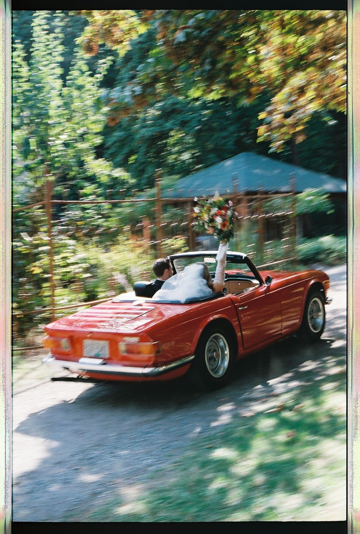 Newlyweds driving away in a classic red convertible sports car decorated with bridal bouquet.
