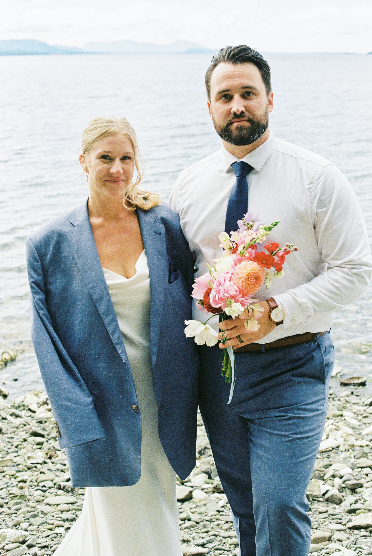 Bride wearing grooms blue blazer holding colorful bouquet with bearded groom by the lake.