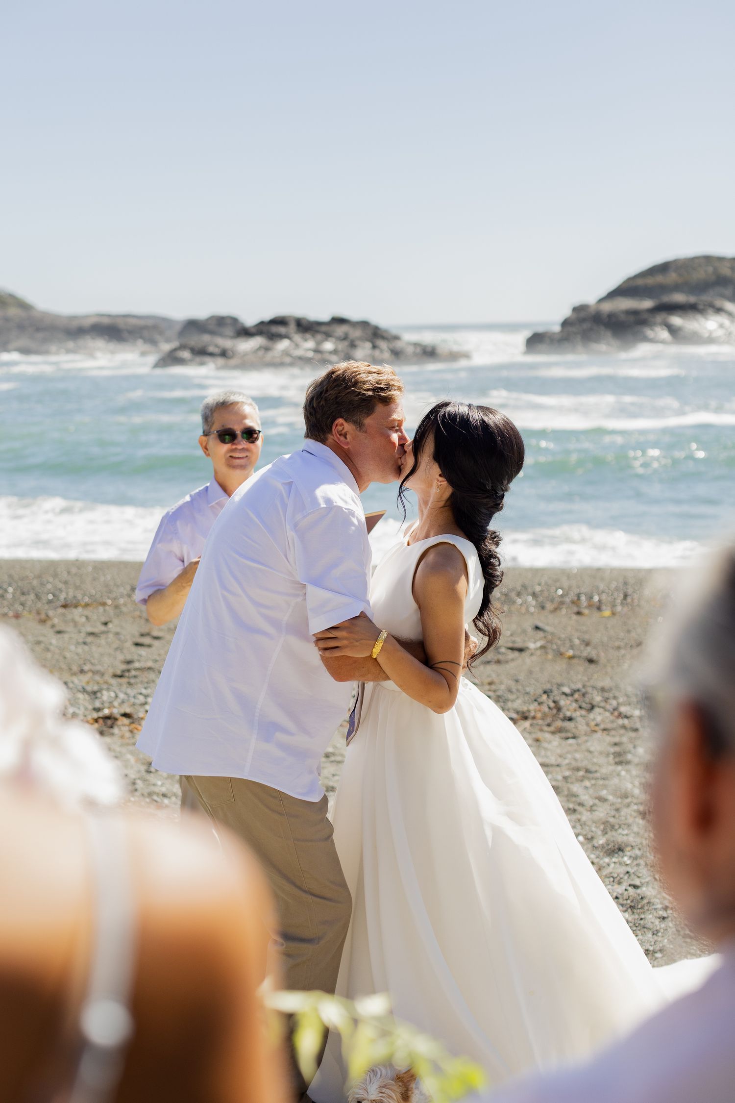 Groom kisses bride during beach wedding ceremony with crashing ocean waves and rocky coastline behind them.
