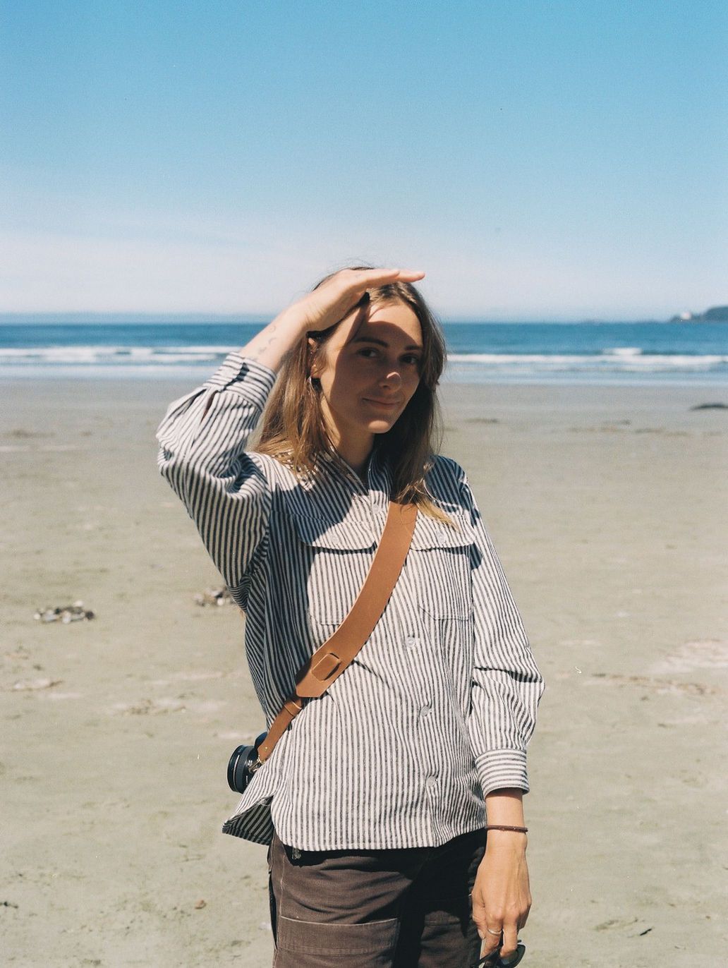 Blonde woman in striped shirt shades her eyes while standing on a sunny sandy beach.