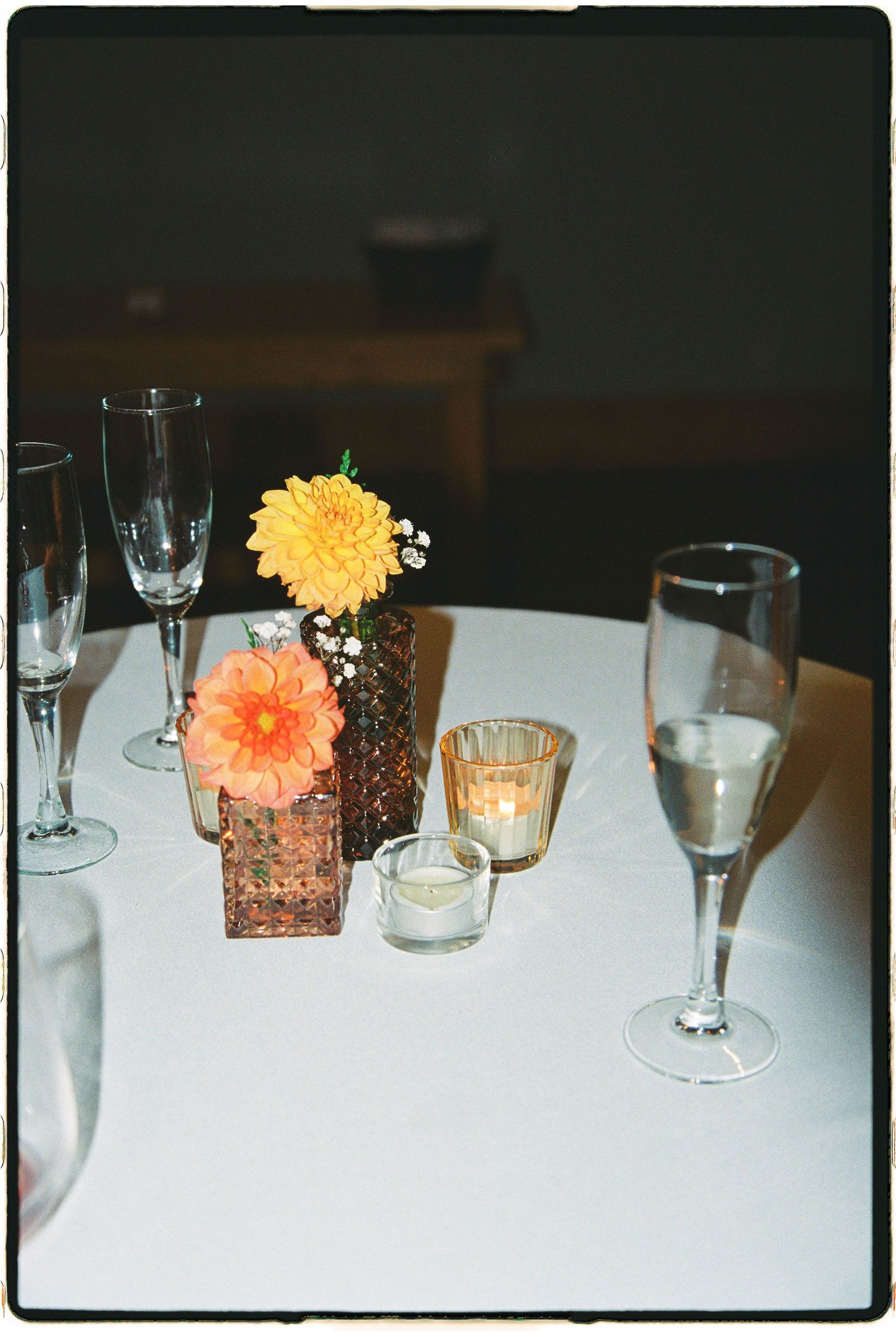 Elegant wedding table centerpiece with orange dahlia flowers in copper vases, candles, and champagne flutes on white linen.