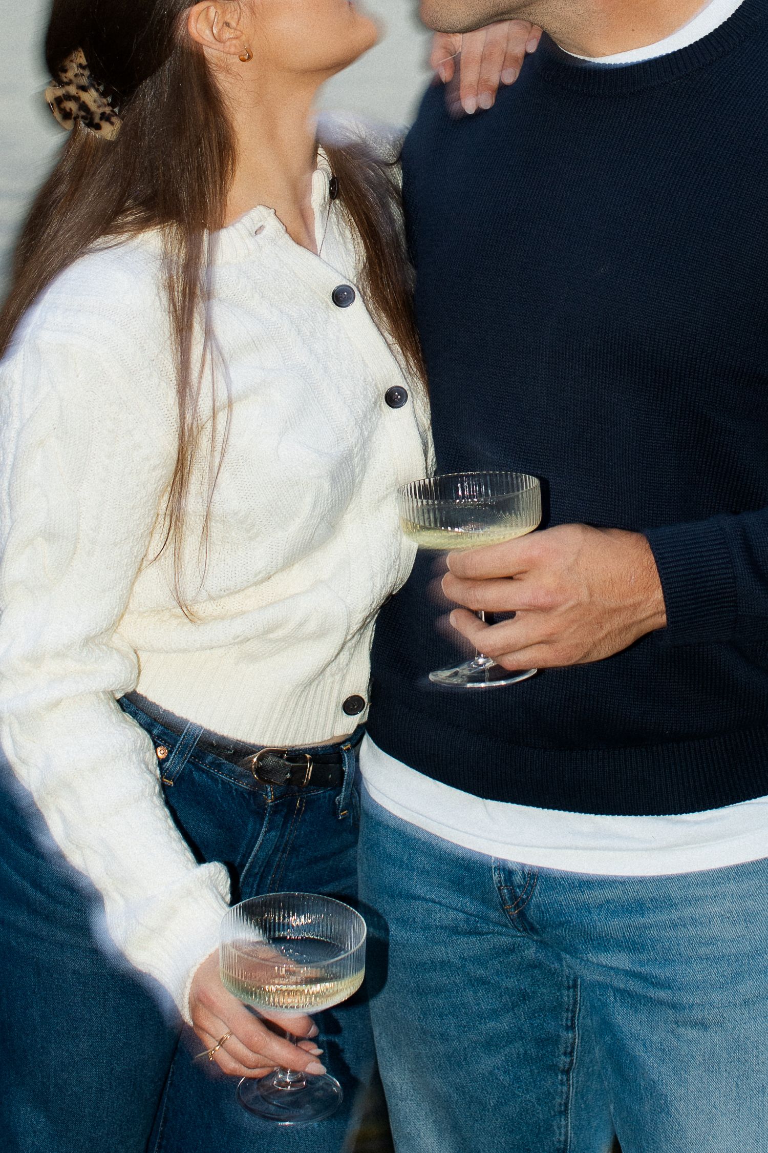 Couple holding champagne coupe glasses together, woman in white cardigan and jeans.
