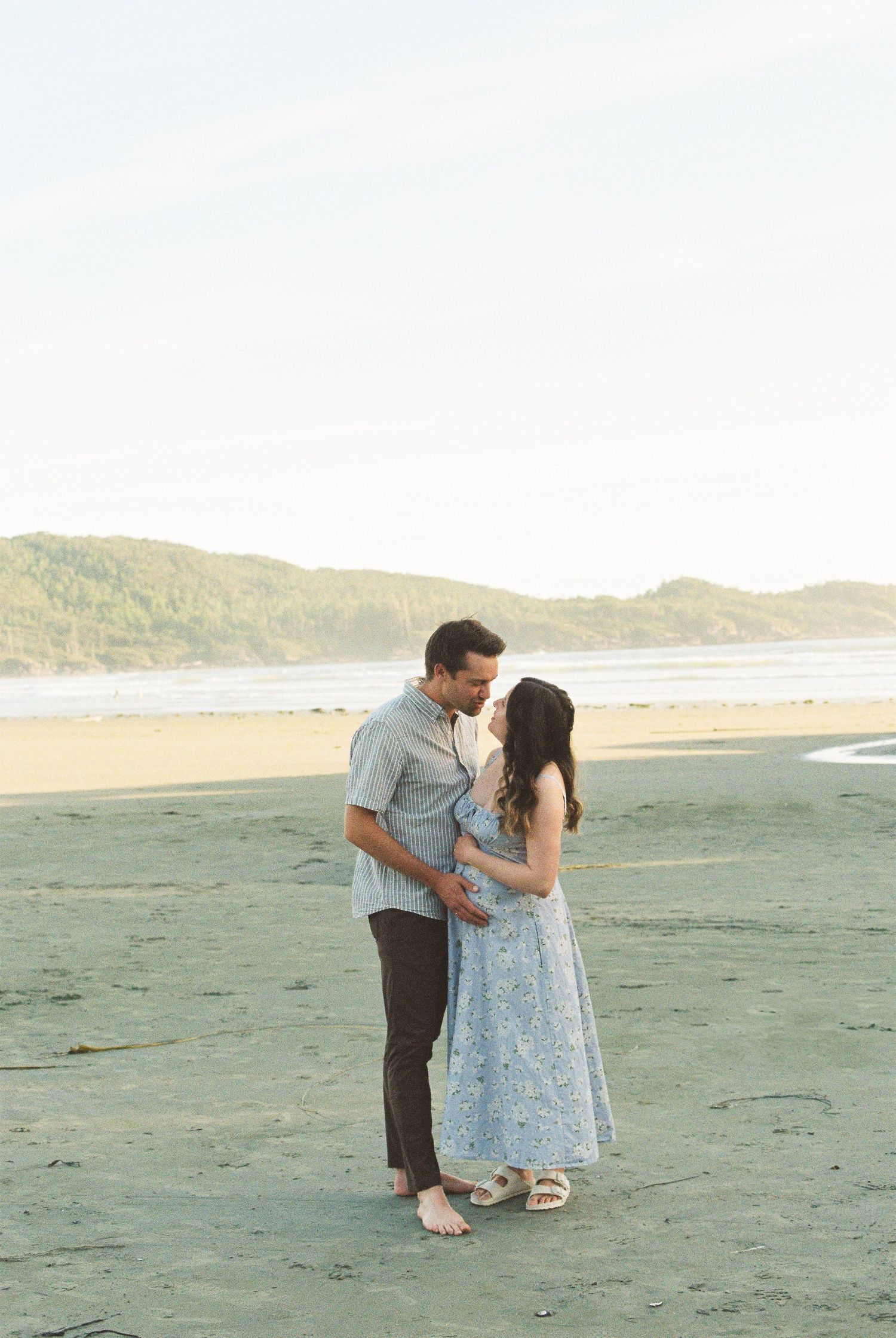 Couple sharing an intimate moment on a sandy beach, smiling at each other with coastal hills in background.