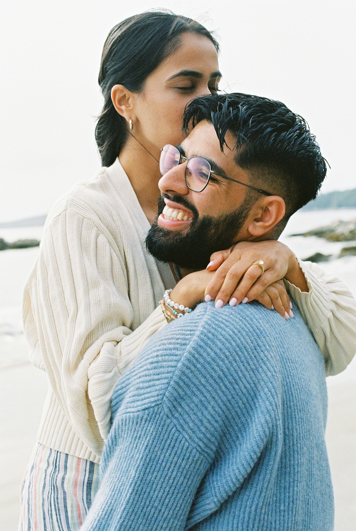 Happy couple embracing on a beach, woman hugging man from behind as he smiles wearing glasses and blue knit sweater.