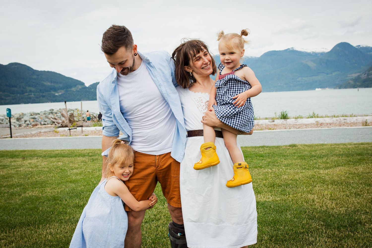 Family of four with two toddlers posing on grass near a scenic mountain lake.