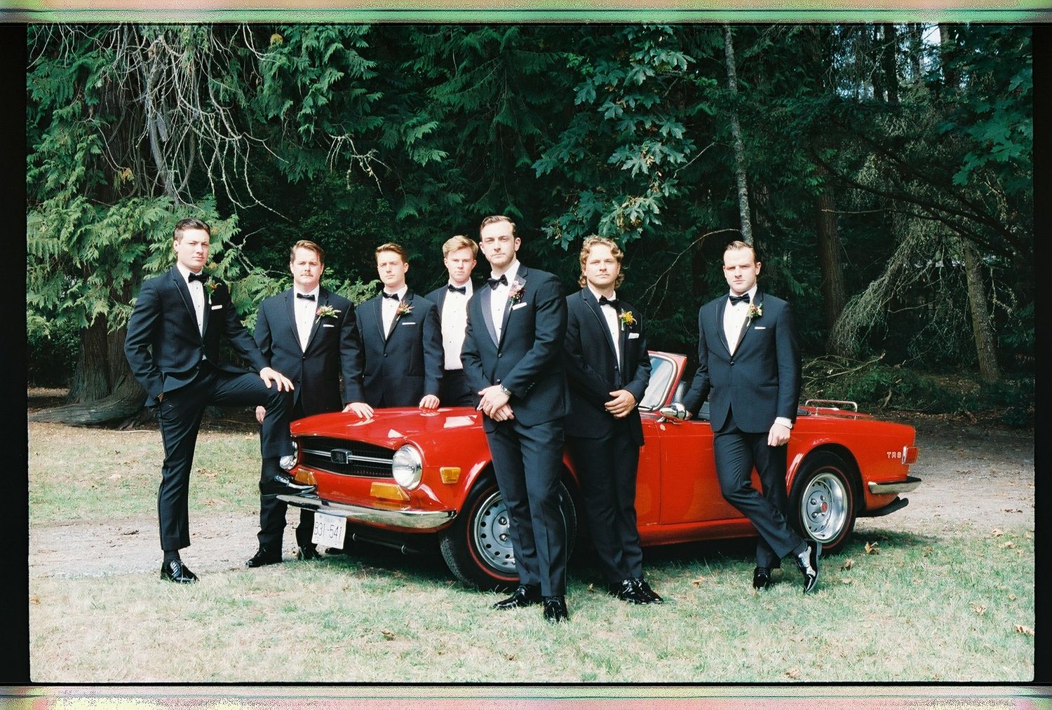 Groom and groomsmen in matching black tuxedos pose beside a vintage red convertible outdoors.
