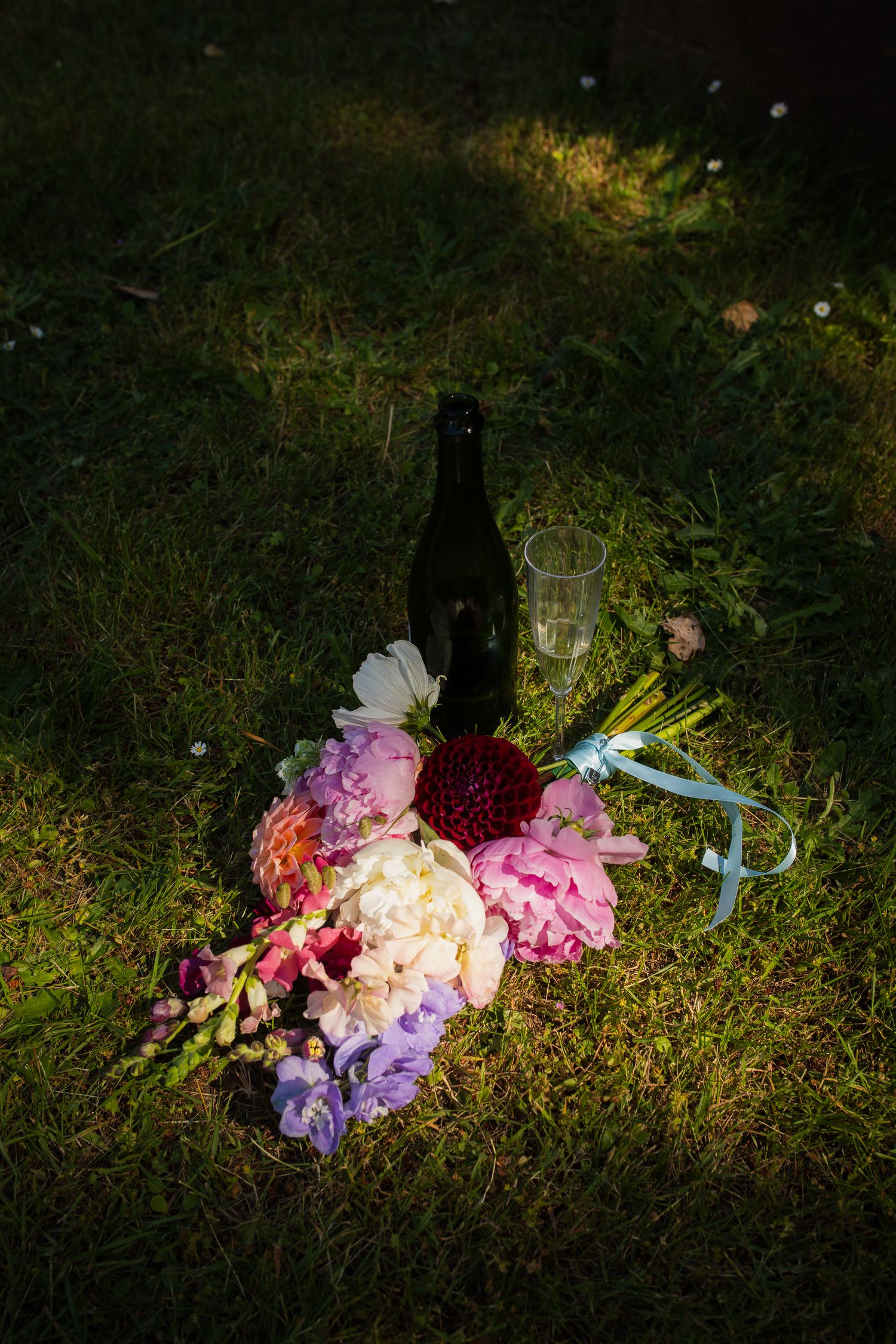 Colorful wedding bouquet with peonies beside a champagne bottle on grass in soft light.