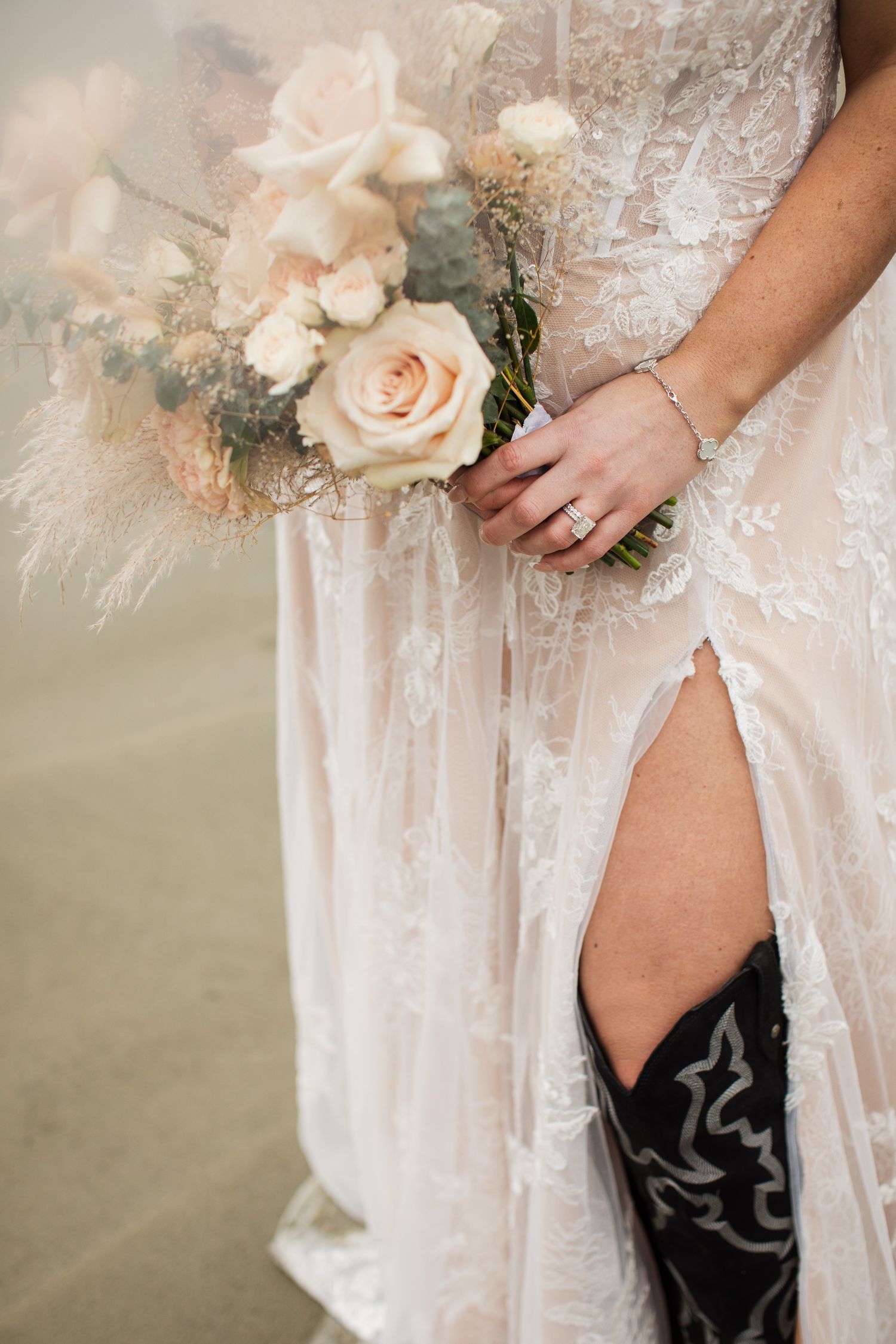 Close-up of bride holding blush rose bouquet in lace wedding gown with cowboy boots on beach.