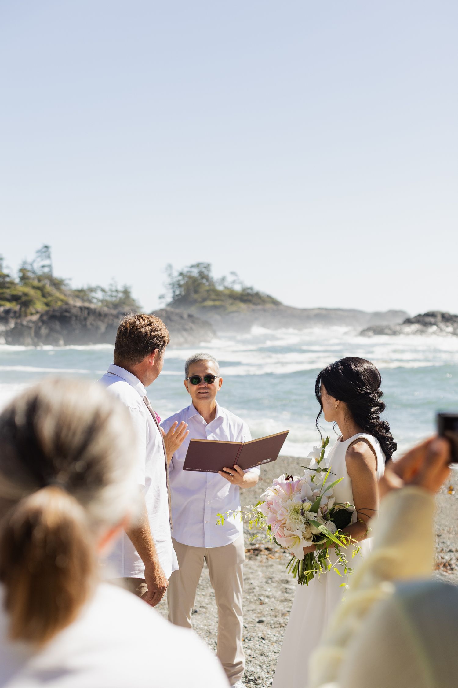 Officiant reads vows to couple during intimate beach wedding ceremony with ocean waves in background.