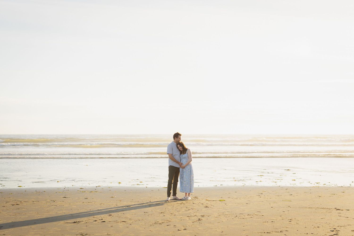 Couple embracing on a wide open sandy beach with calm ocean waters and bright sky in the background.