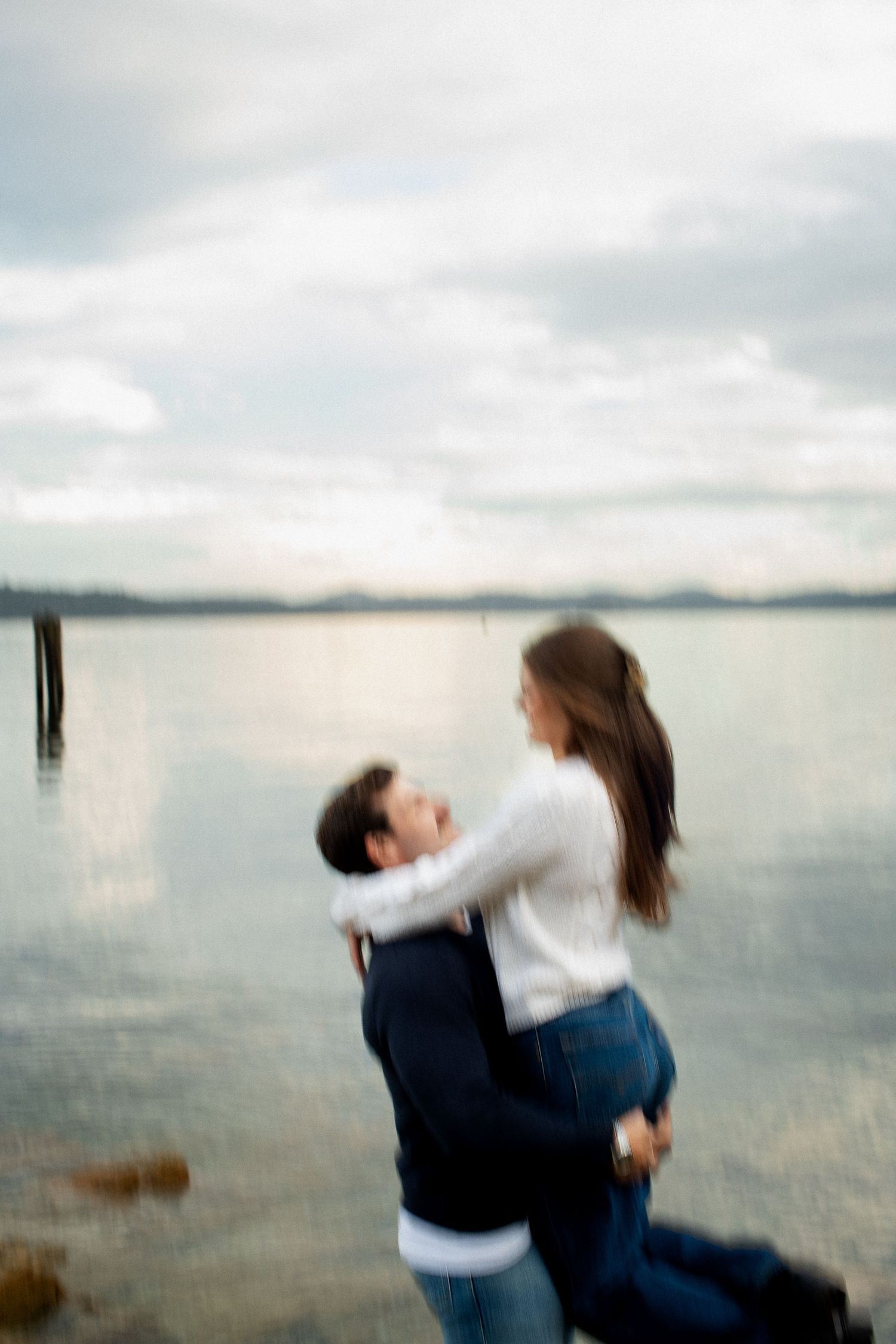 Couple embracing romantically by calm lake shore under cloudy sky, motion blur effect.