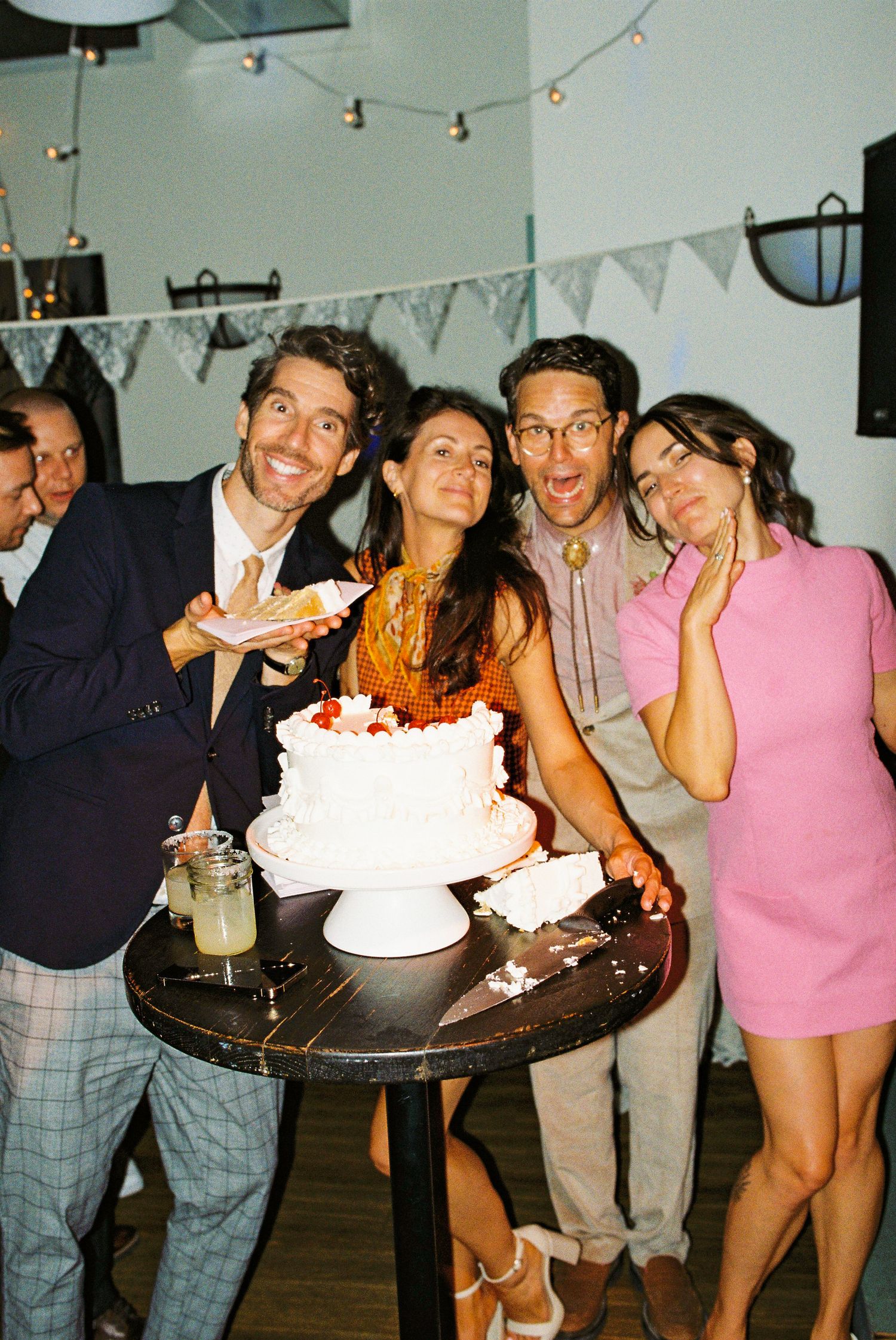 Two couples laugh and pose around a white wedding cake at a festive indoor reception celebration.