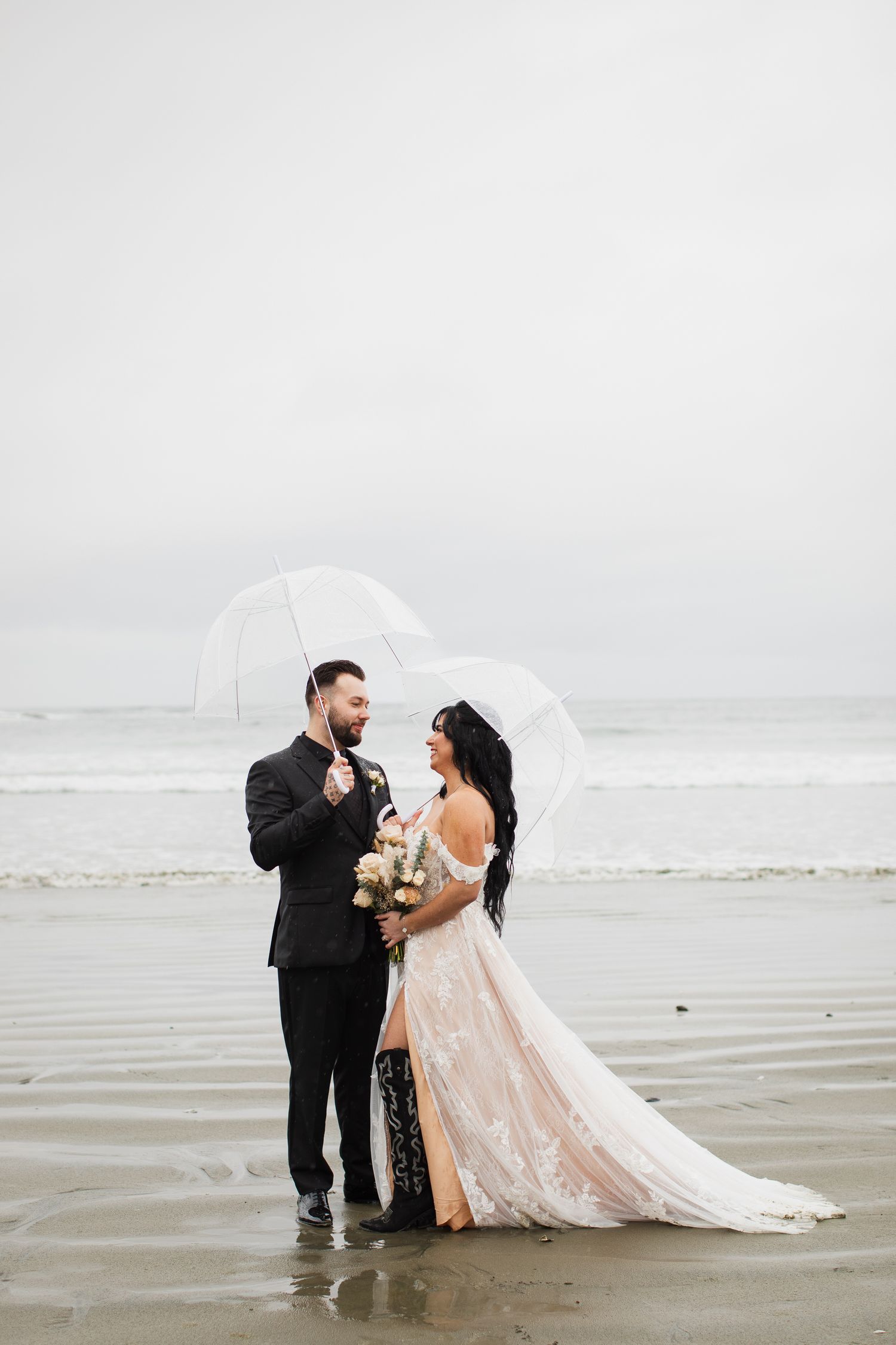 Bride and groom stand under clear umbrella on misty beach, bride holding bouquet of neutral flowers.