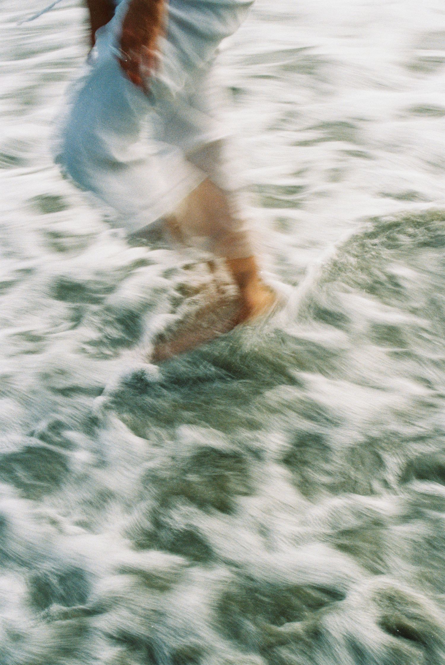 Blurred motion shot of barefoot person in white dress running through ocean waves on beach.