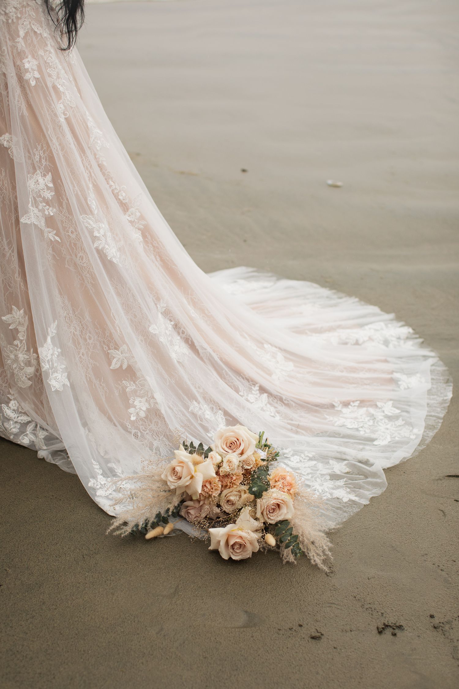 Bride's blush lace wedding gown train spreads across wet sand beside a peach rose bouquet.