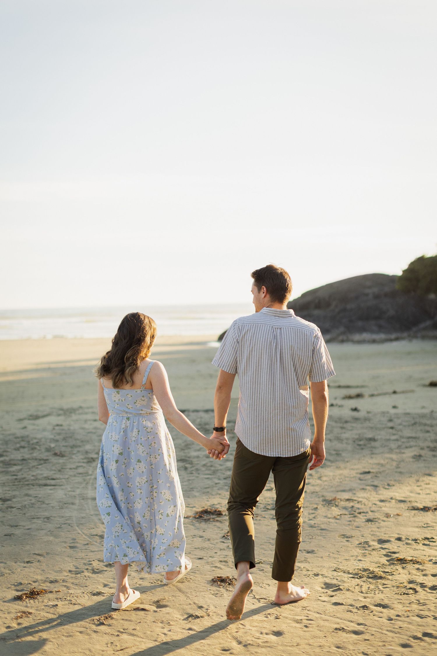 A couple walks hand in hand barefoot along a sunny sandy beach toward the ocean.
