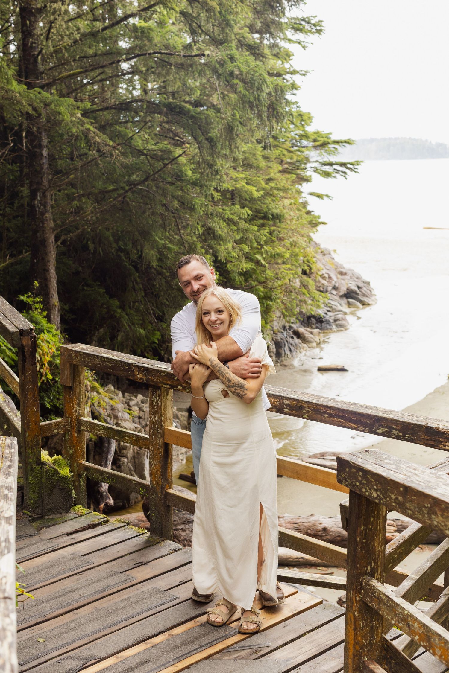 Couple embracing on a wooden deck overlooking a rocky, tree-lined coastal shoreline.