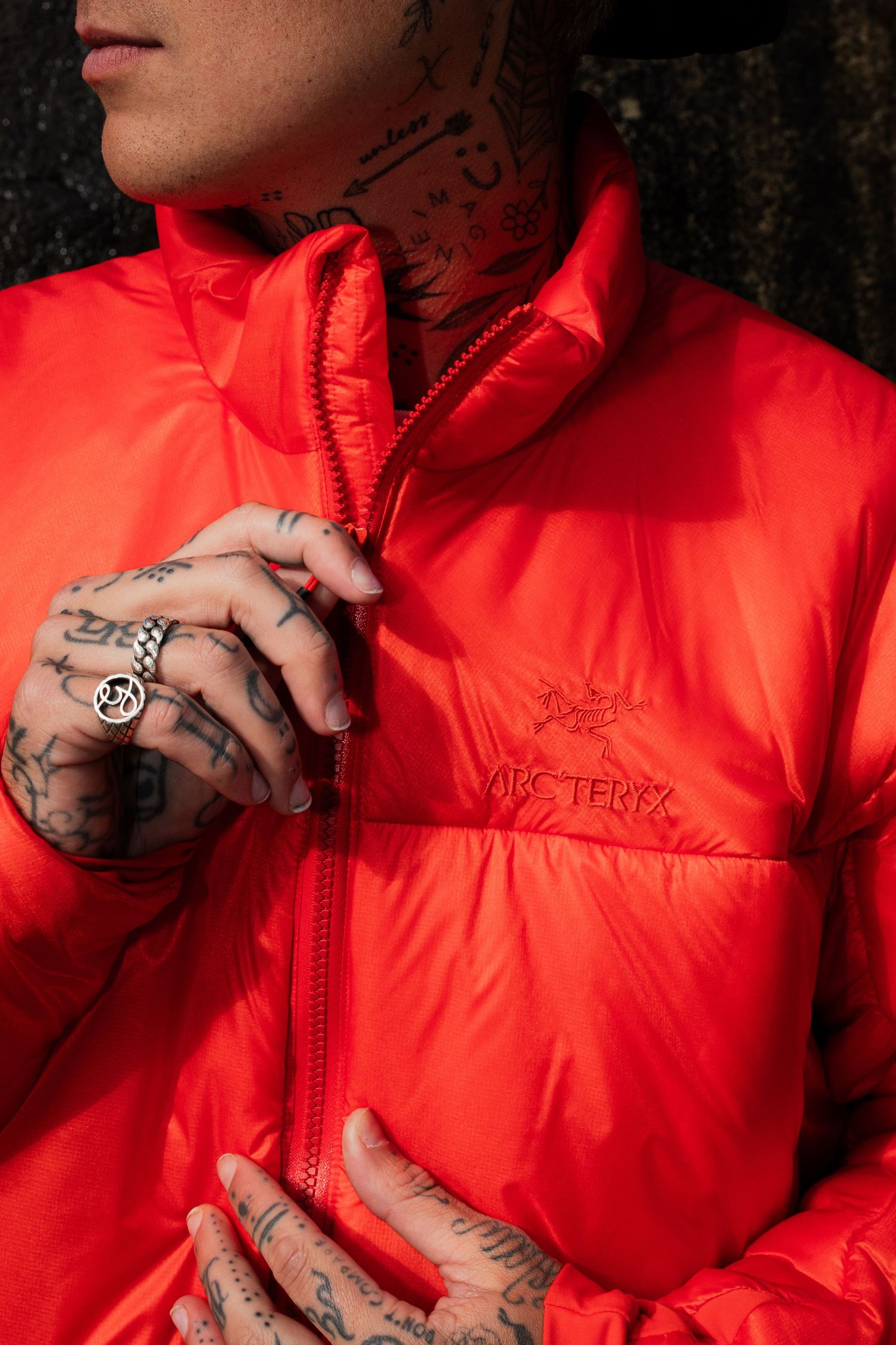 Tattooed person wearing a bright red Arc'teryx puffer jacket zipping it up outdoors.
