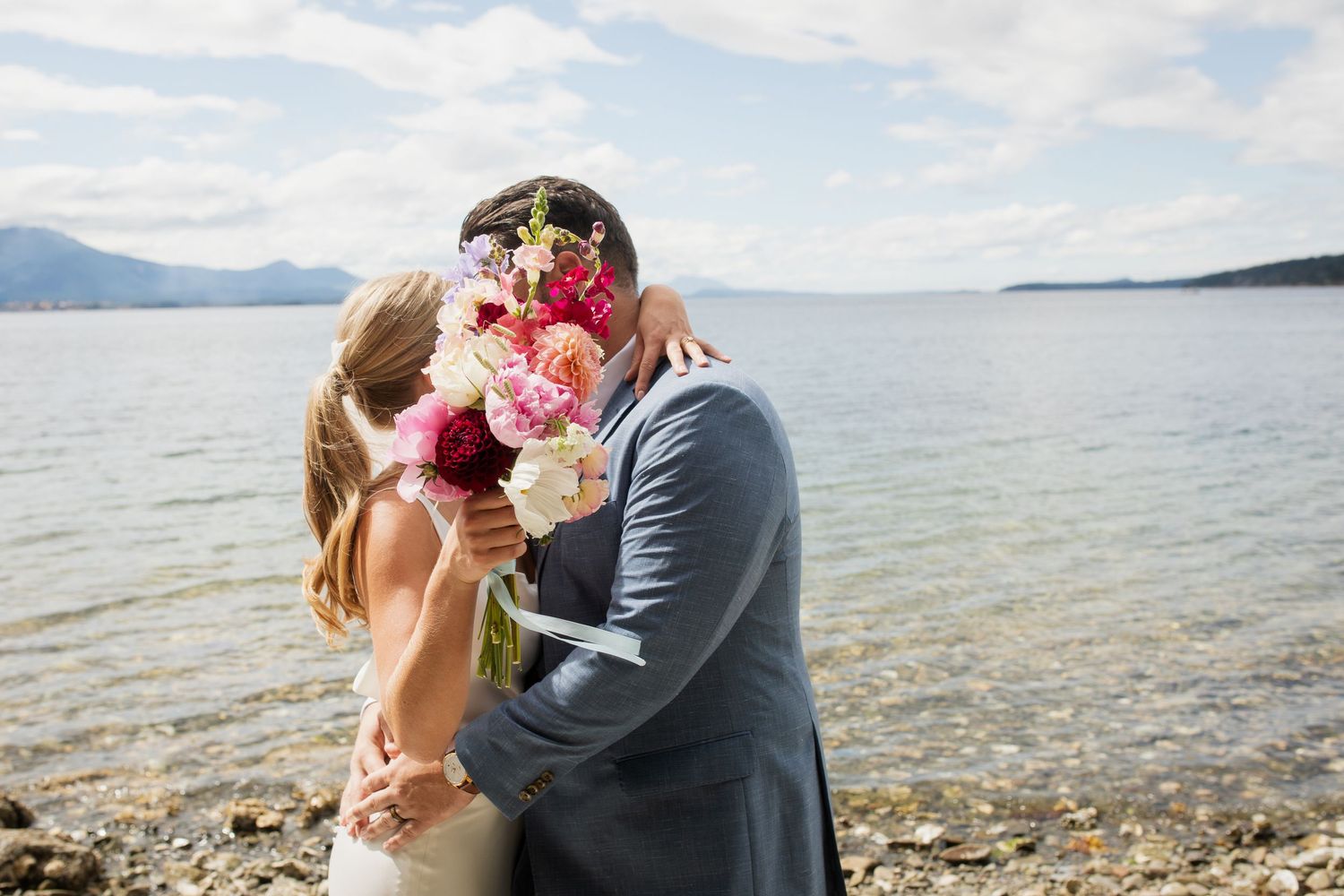 Bride and groom kiss behind colorful pink and white bouquet beside a scenic mountain lake.