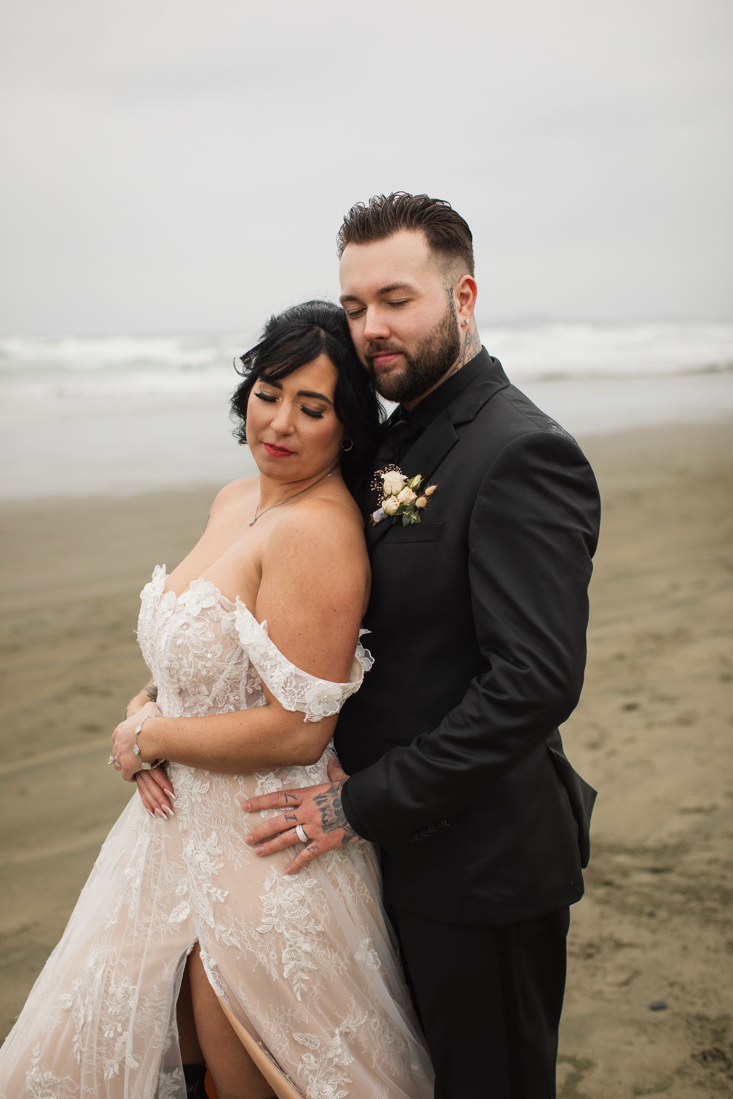 Bride and groom embrace on a foggy beach, she in lace off-shoulder gown, he in black suit with boutonniere.