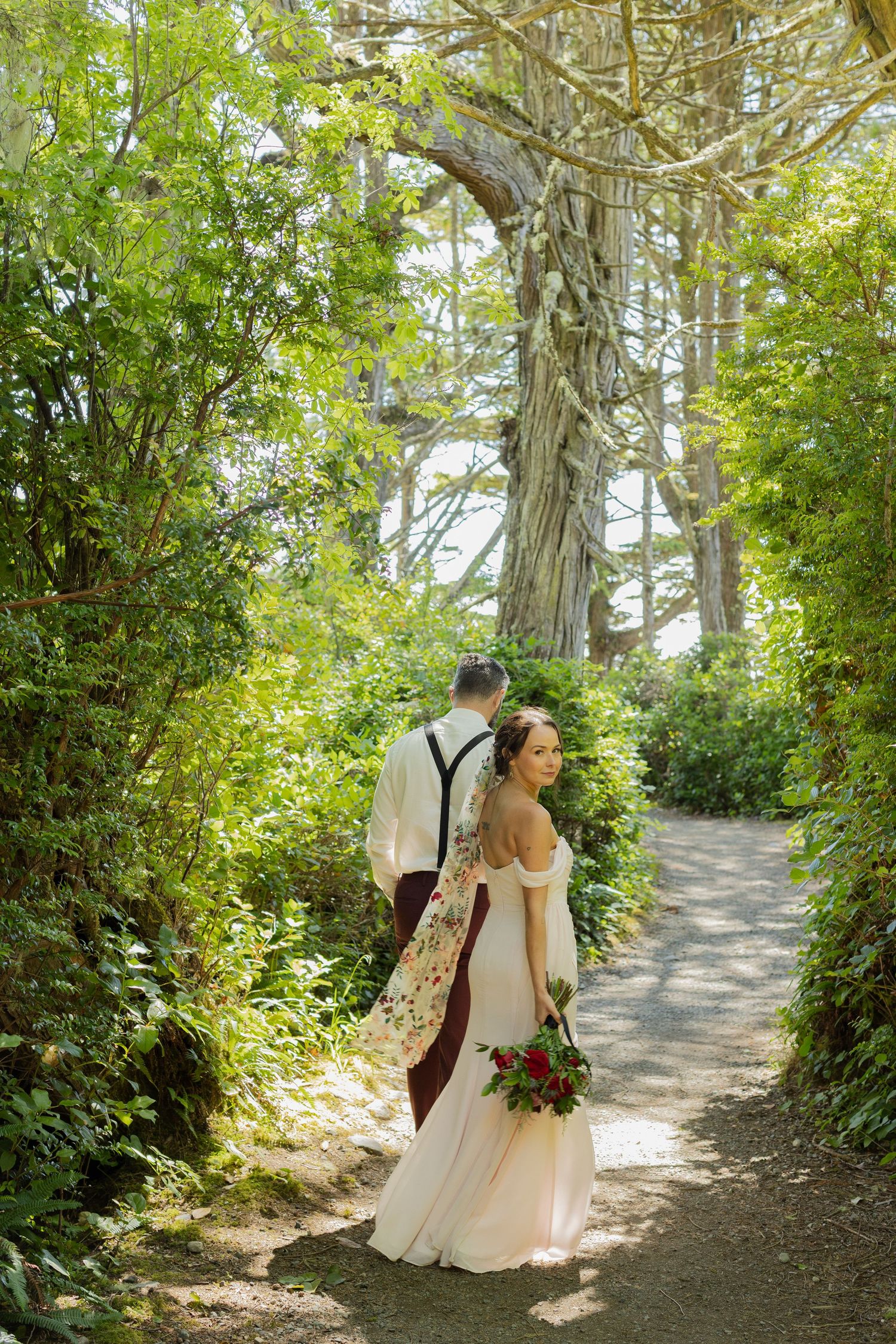 Bride and groom walk hand-in-hand along a sunlit forest path with lush green trees.