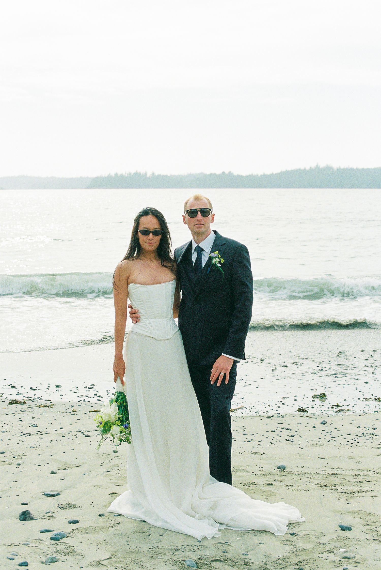 Bride and groom in sunglasses posing together on sandy beach with calm ocean waters behind them.