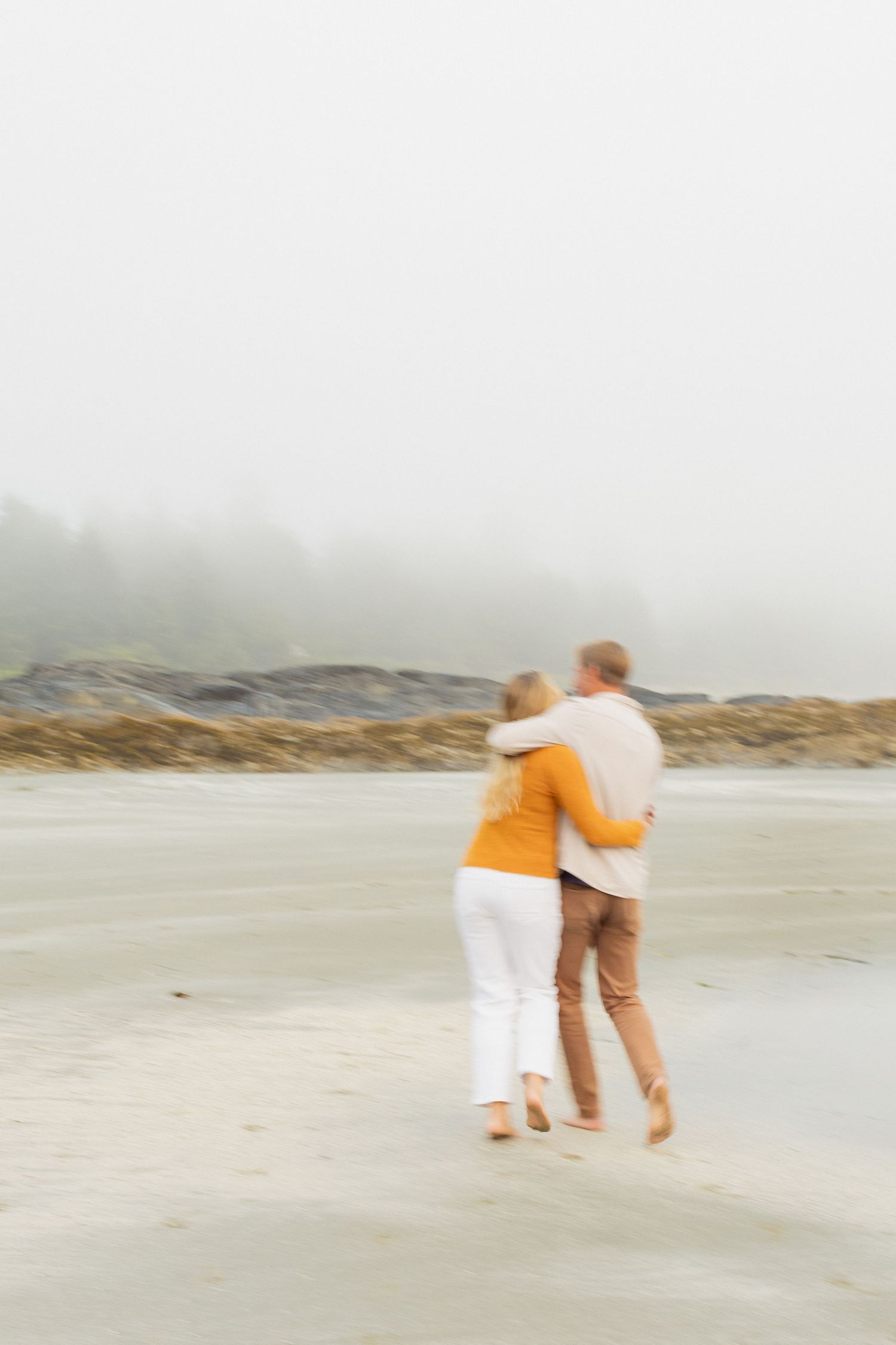 Couple embracing and walking together on misty sandy beach with rocky coastline in background.