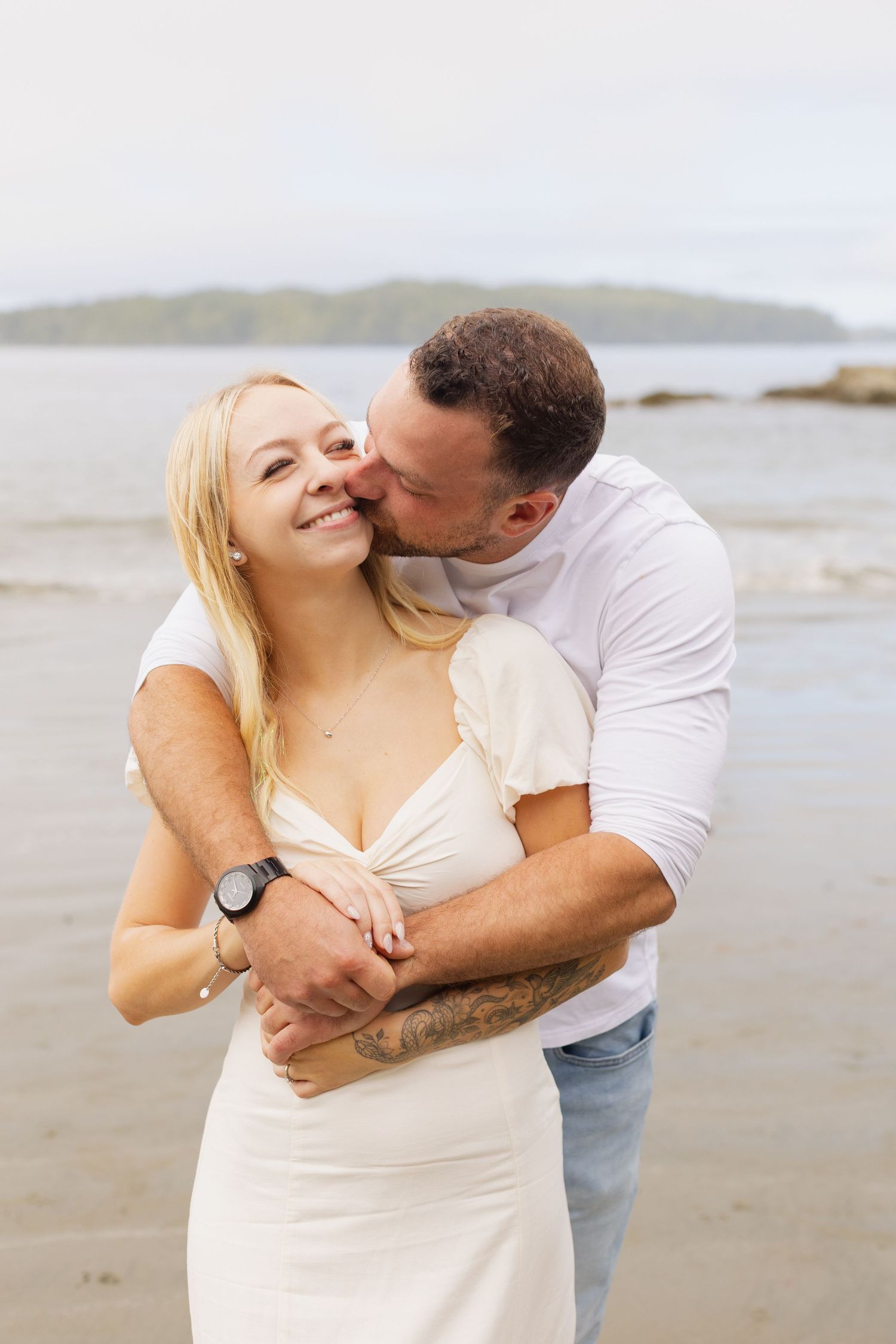 Man kissing blonde woman on cheek while embracing her on a sandy beach shoreline.