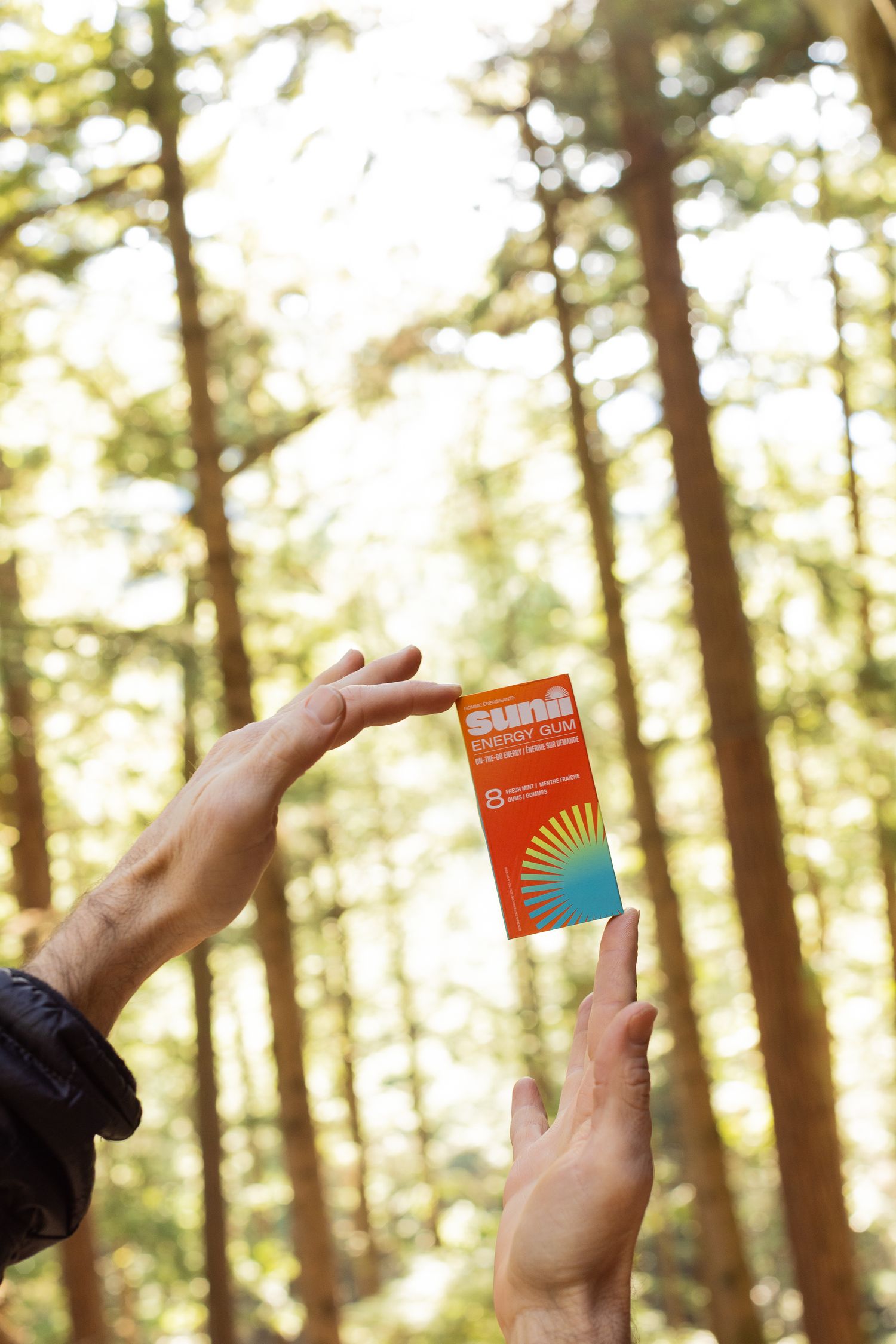Two hands holding a Sunsi sunscreen box up against tall sunlit forest trees with bokeh background.