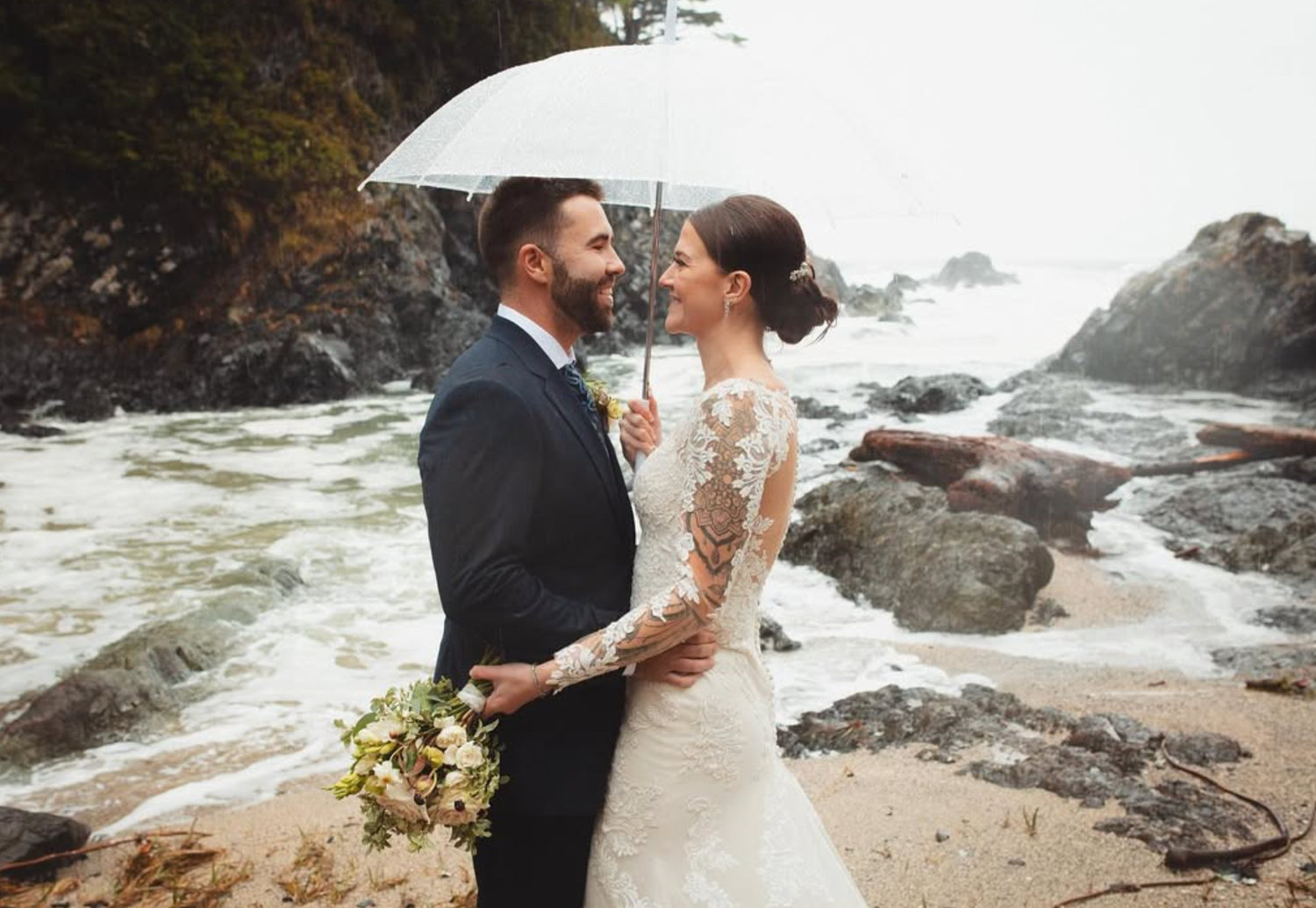 Bride and groom share romantic moment under clear umbrella on rocky beach in the rain.