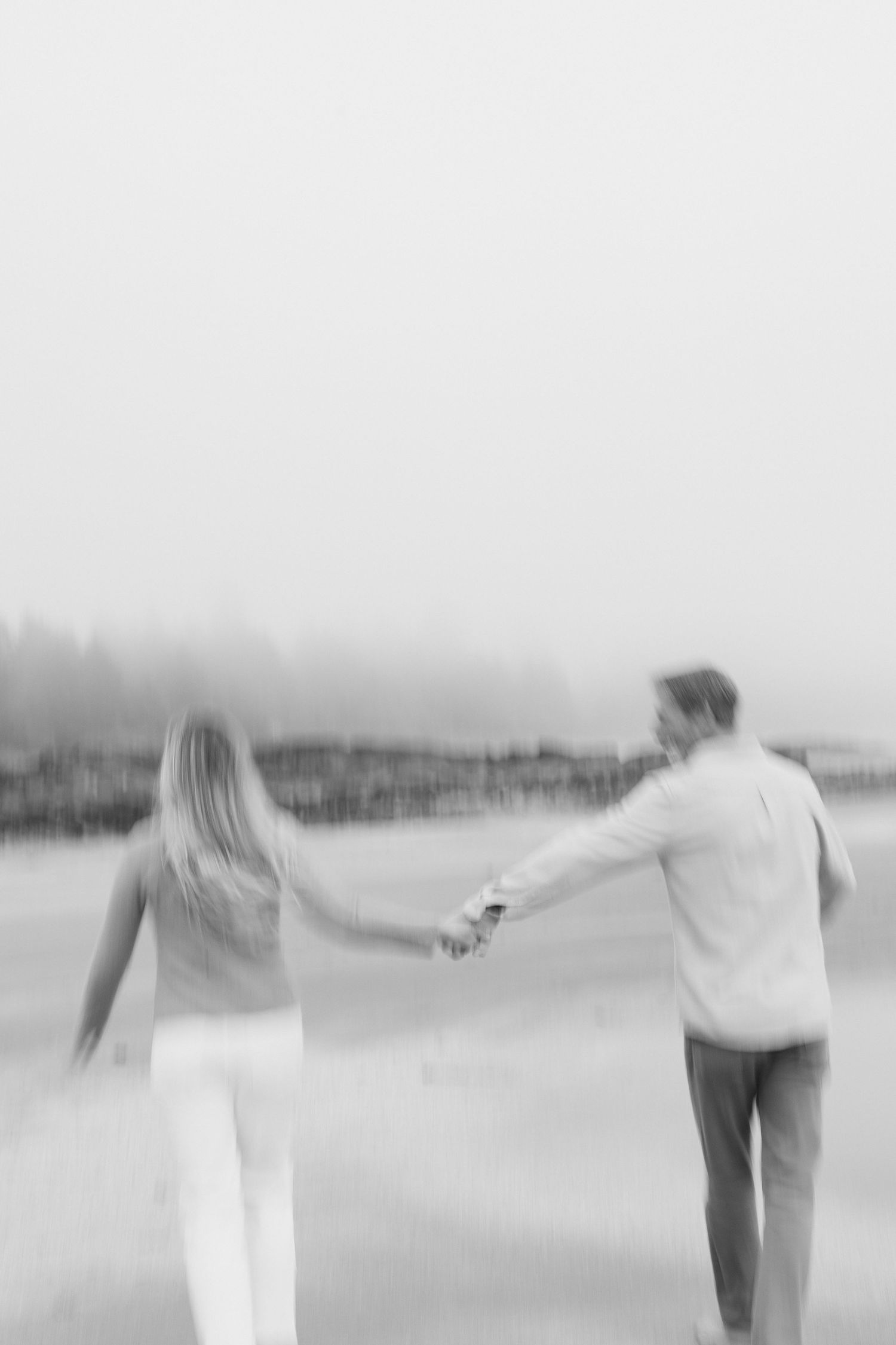 Blurred black and white artistic photo of couple holding hands walking on foggy beach.