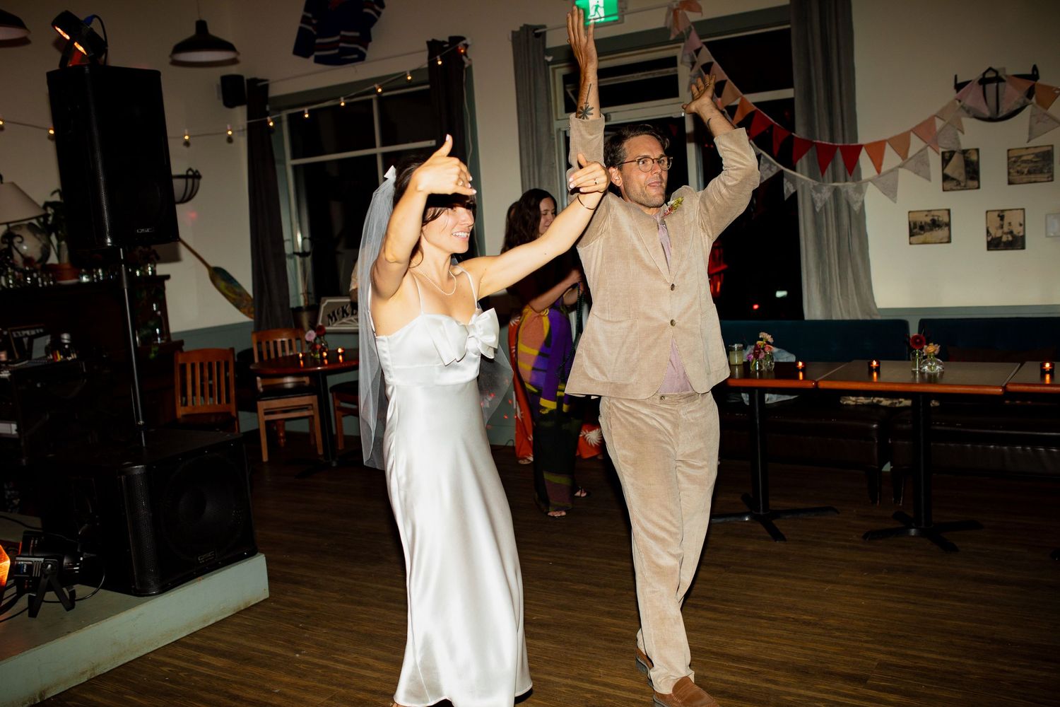 Bride in white slip dress and groom dancing energetically together at indoor wedding reception.