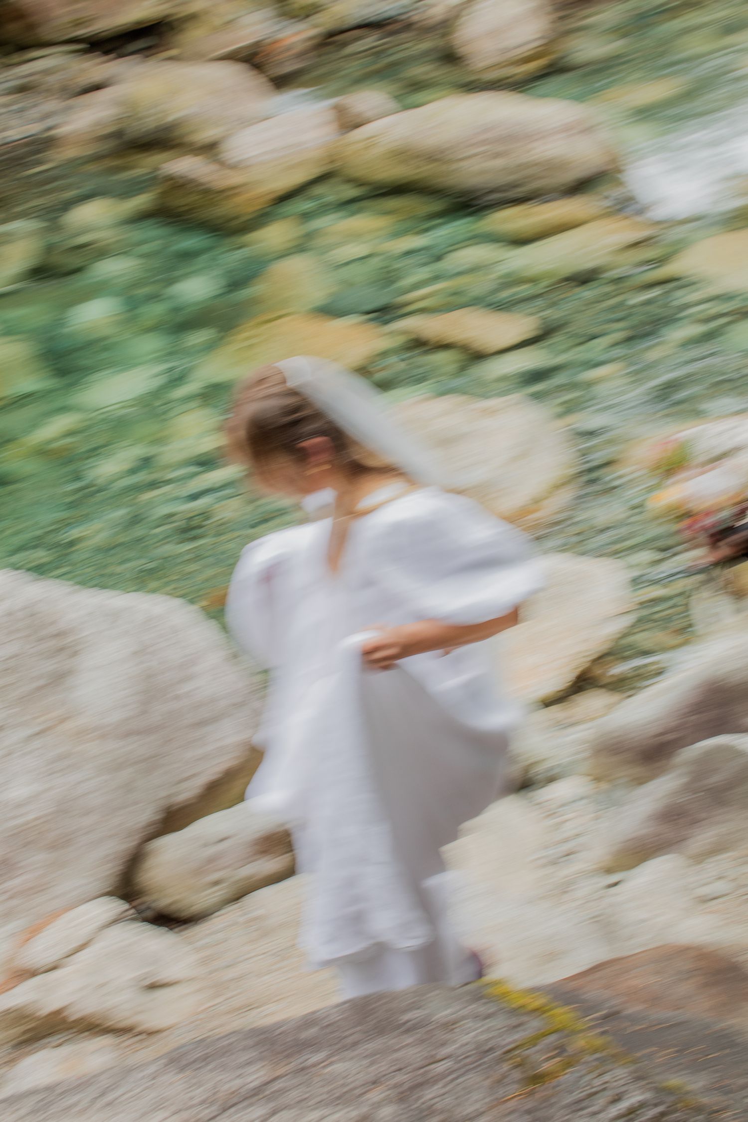 Artistic blurred motion photo of bride in white dress walking among rocky landscape.