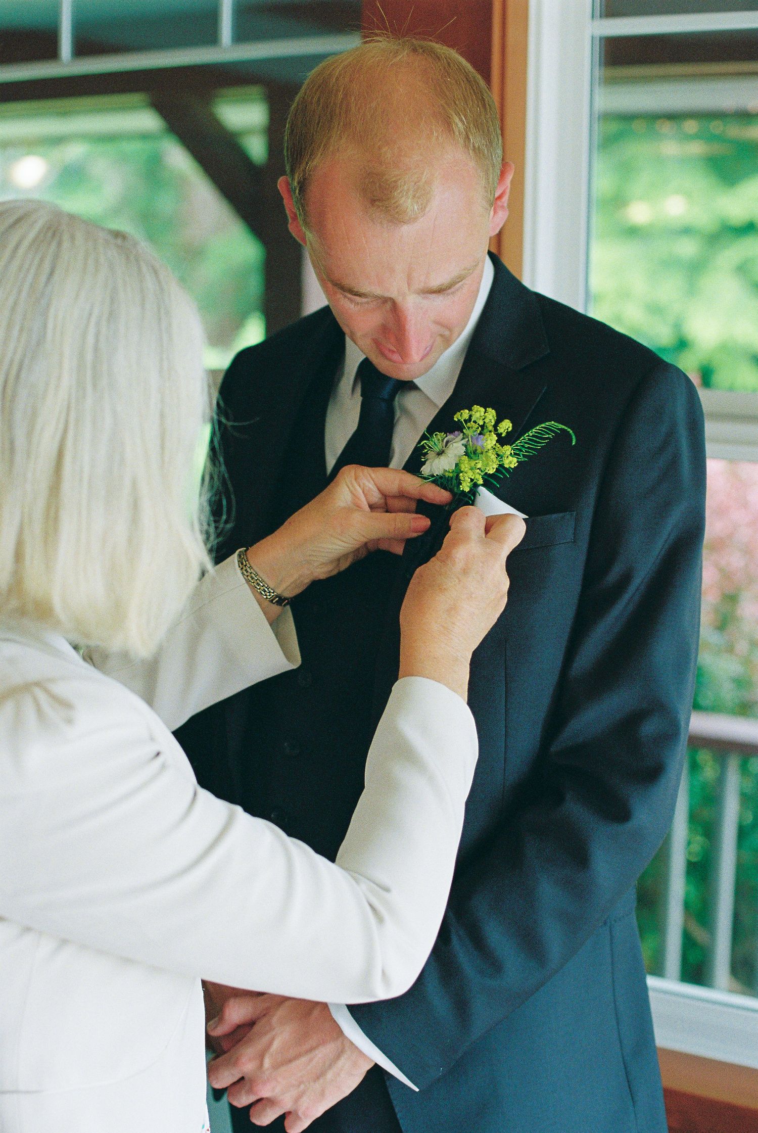 Mother pins a green boutonniere onto groom's navy suit lapel before wedding ceremony indoors.
