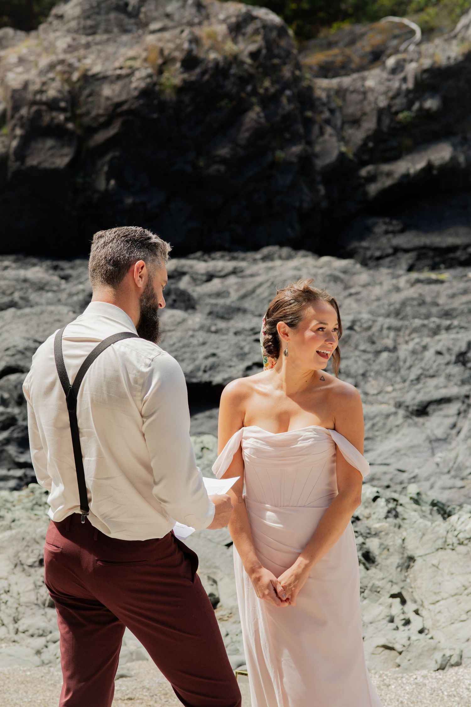 Bearded groom in suspenders reads vows to smiling bride in blush dress at rocky beach.