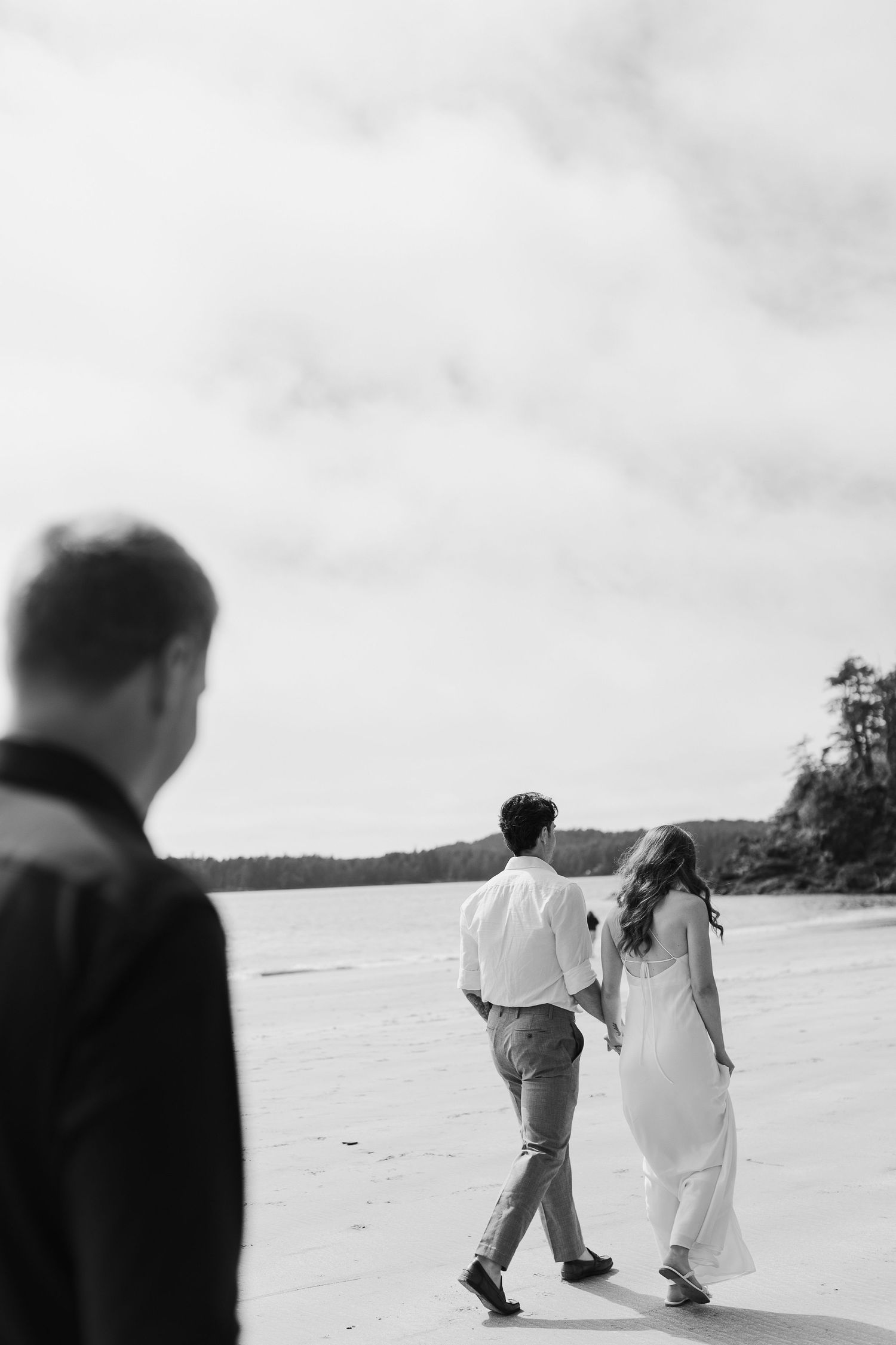 Black and white photo of bride and groom walking hand in hand on beach.