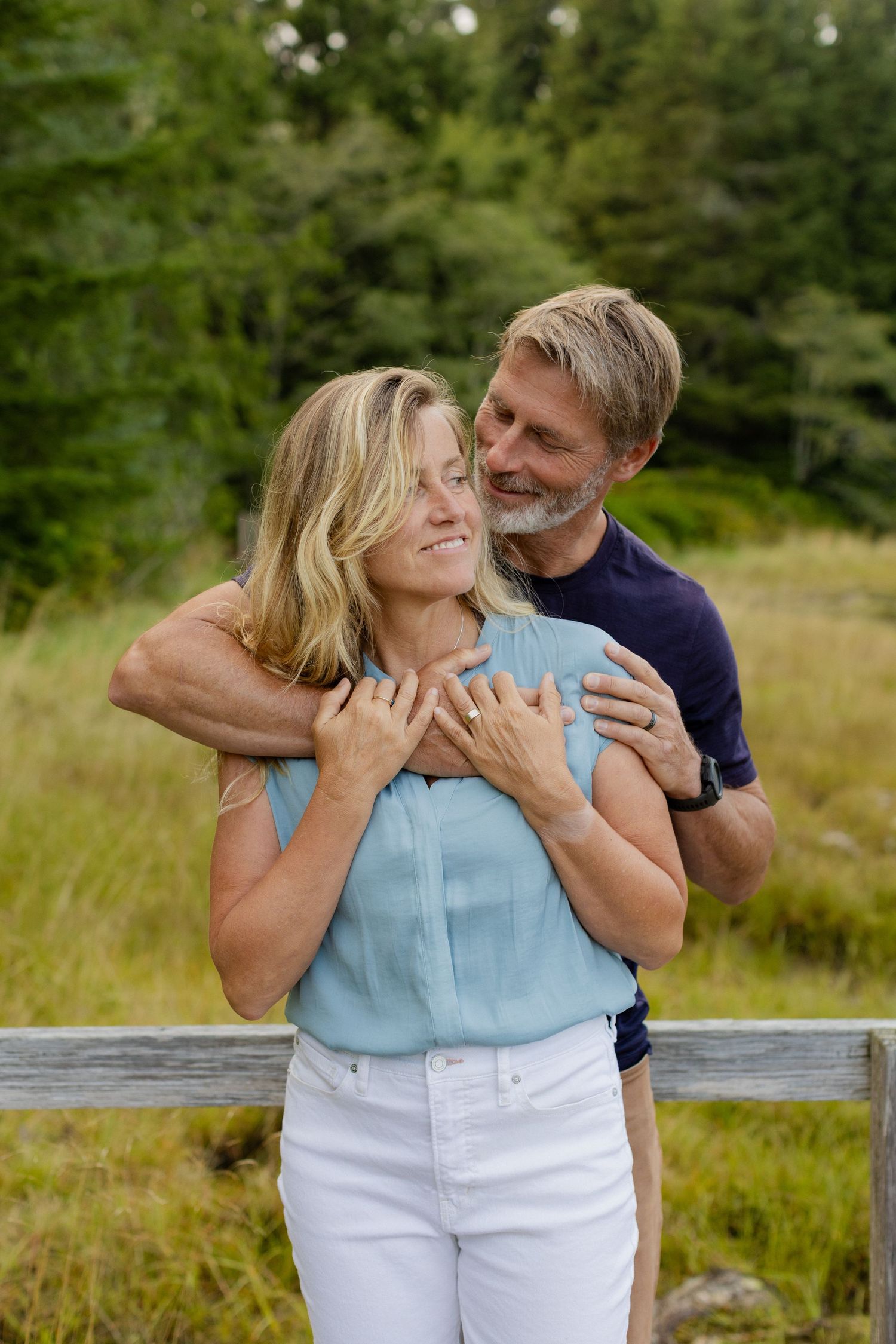 A man embraces a smiling blonde woman from behind near a wooden fence surrounded by lush greenery.