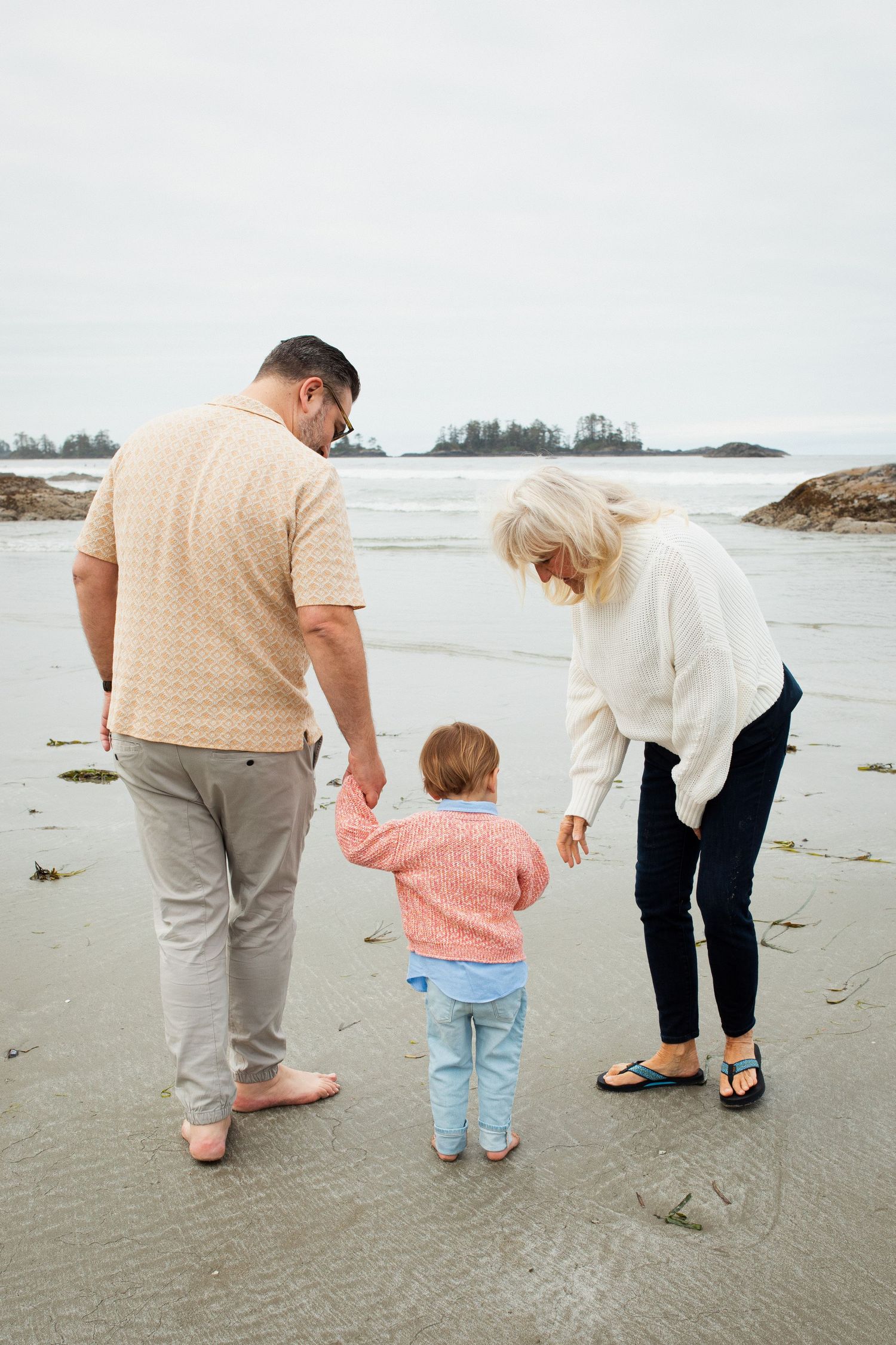Grandparents hold hands with a toddler while standing at the ocean's edge on a sandy beach.
