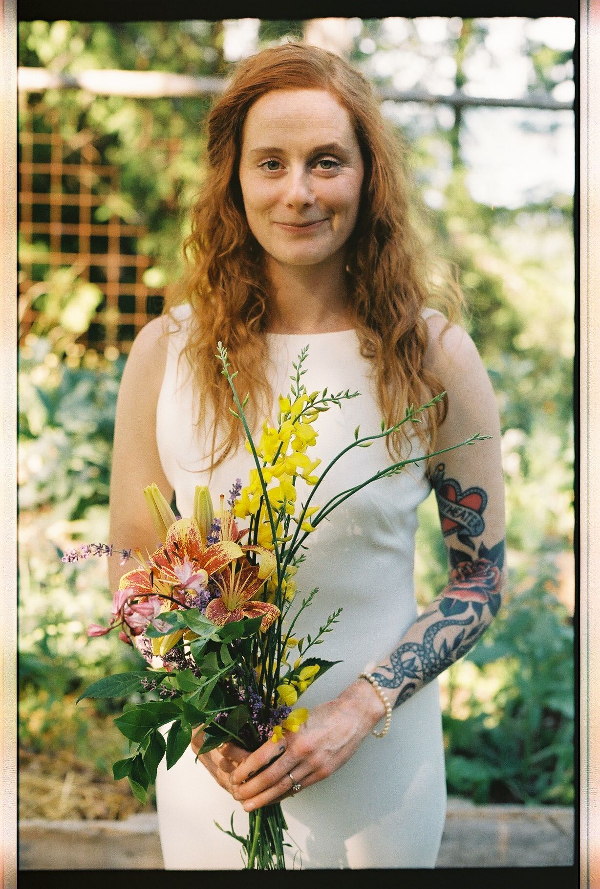 Red-haired bride with tattoo sleeve holding colorful wildflower bouquet at outdoor garden wedding.