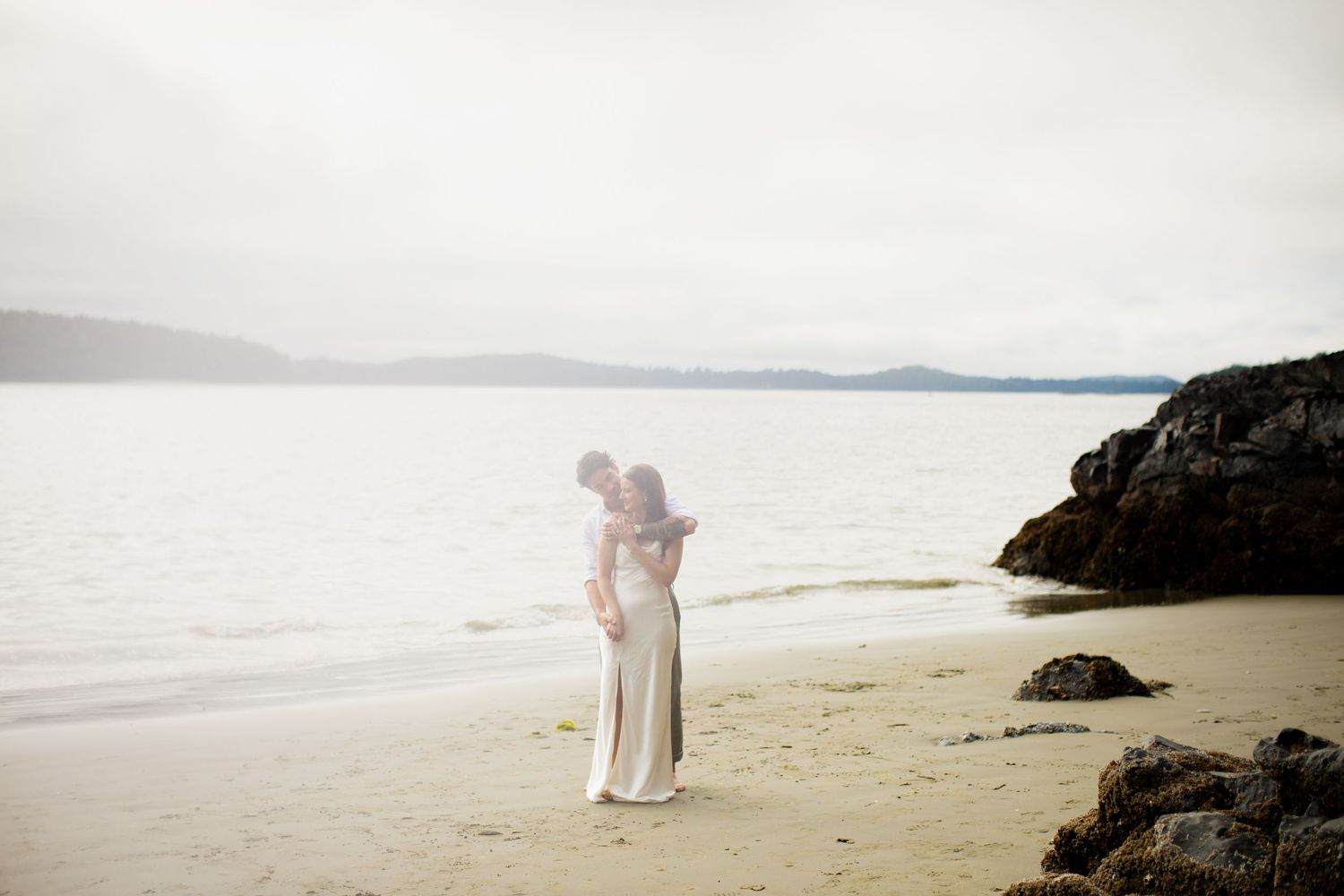 Couple embracing on a serene beach with calm water and misty mountains in the background.