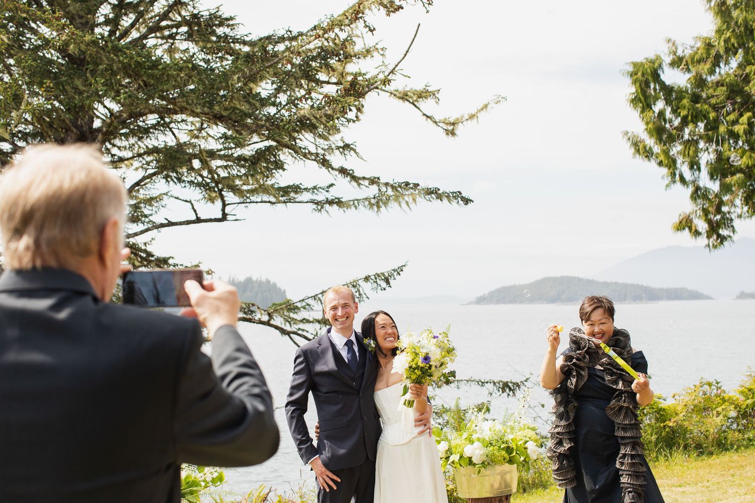 Wedding couple posing for photos on grassy lawn with pine tree and ocean backdrop while guests celebrate.